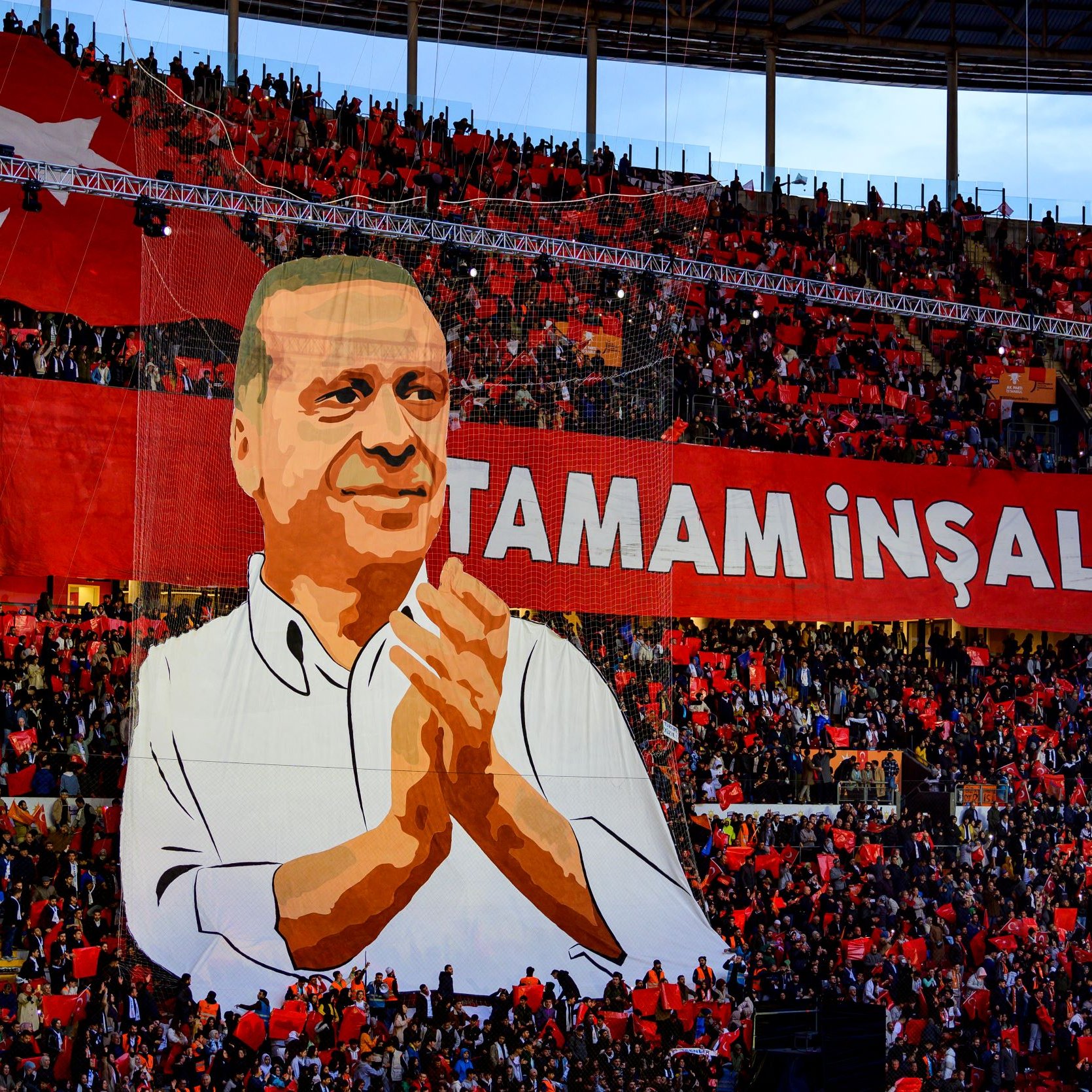 L'image montre une grande scène dans un stade rempli de supporters. Au centre, il y a une immense illustration d'un homme qui semble être un leader ou un personnage politique, probablement Erdoğan, qui applaudit. En arrière-plan, une banderole rouge avec des inscriptions en lettres blanches affiche "TAMAM İNŞALLAH". Les spectateurs tiennent des drapeaux rouges, créant une atmosphère colorée et festive. L'ensemble évoque une manifestation de soutien, probablement lors d'un événement politique.