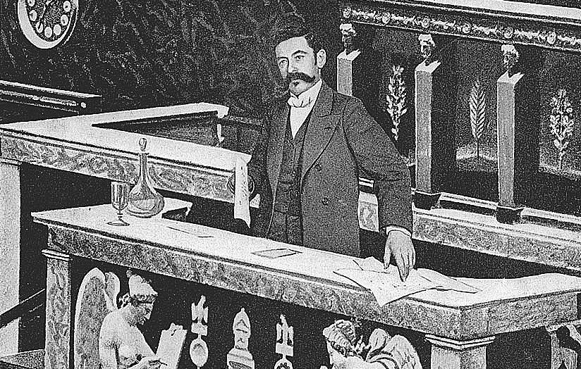 L'image montre un homme bien habillé, debout derrière un bureau ou une table qui semble faire partie d'une institution, probablement un parlement ou une assemblée. Le bureau est décoré avec des motifs ornementaux et des sculptures d'anges de chaque côté. L'homme semble concentré, tenant des documents, et l'ambiance générale est formelle et historique.