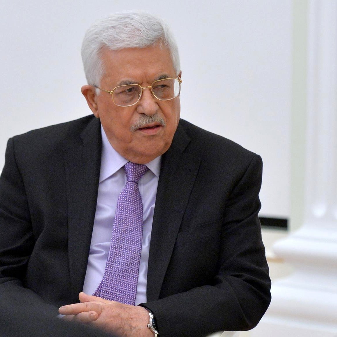 الصورة تظهر رجلًا في ملابس رسمية داكنة، يرتدي نظارات ولديه شعر أبيض. يبدو جالسًا في مكان ذو طابع رسمي، مع خلفية تتضمن جدران بيضاء وملاحظات خفيفة. تعبير وجهه يوحي بأنه في حالة من التفكير أو النقاش.