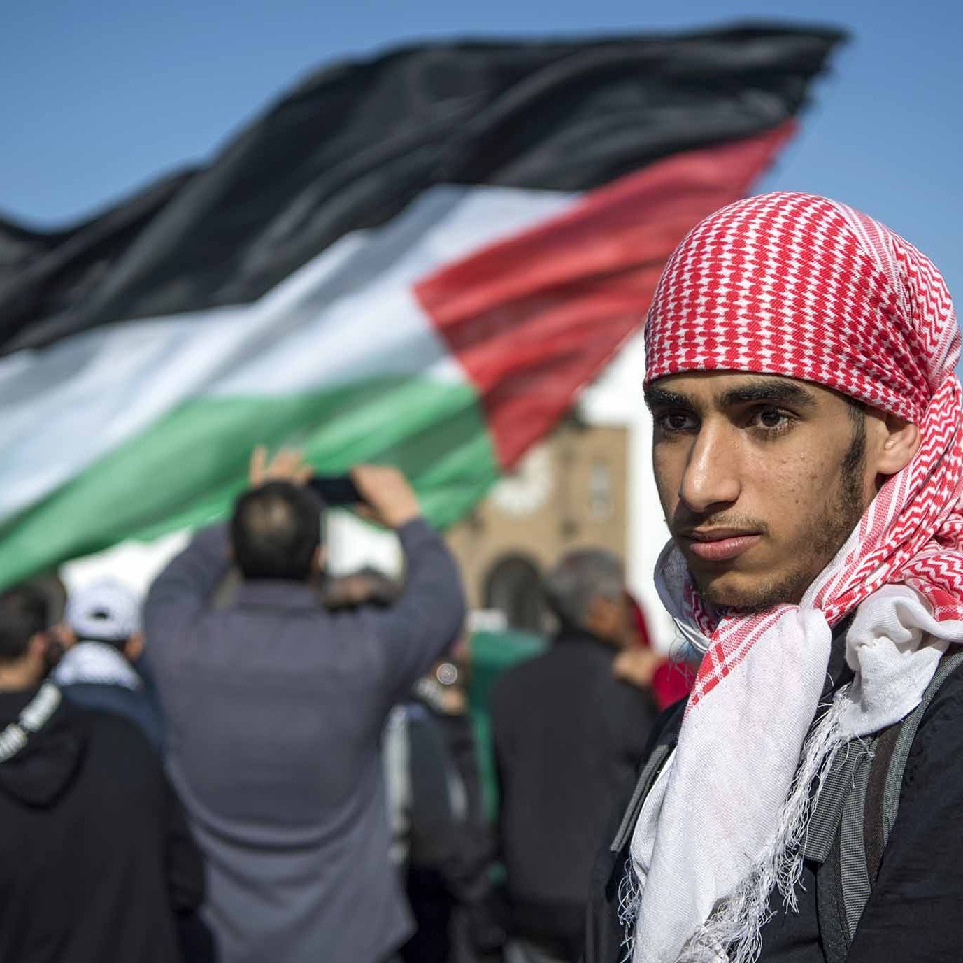 L'image montre un jeune homme portant un keffieh rouge et blanc, se tenant au premier plan. En arrière-plan, une grande foule est visible, avec un drapeau palestinien noir, blanc, vert et rouge qui flotte. L'atmosphère semble être celle d'une manifestation ou d'un rassemblement, où les participants expriment leurs convictions ou leur soutien à une cause. Le jeune homme affiche une expression sérieuse, ce qui ajoute à l'intensité de la scène.