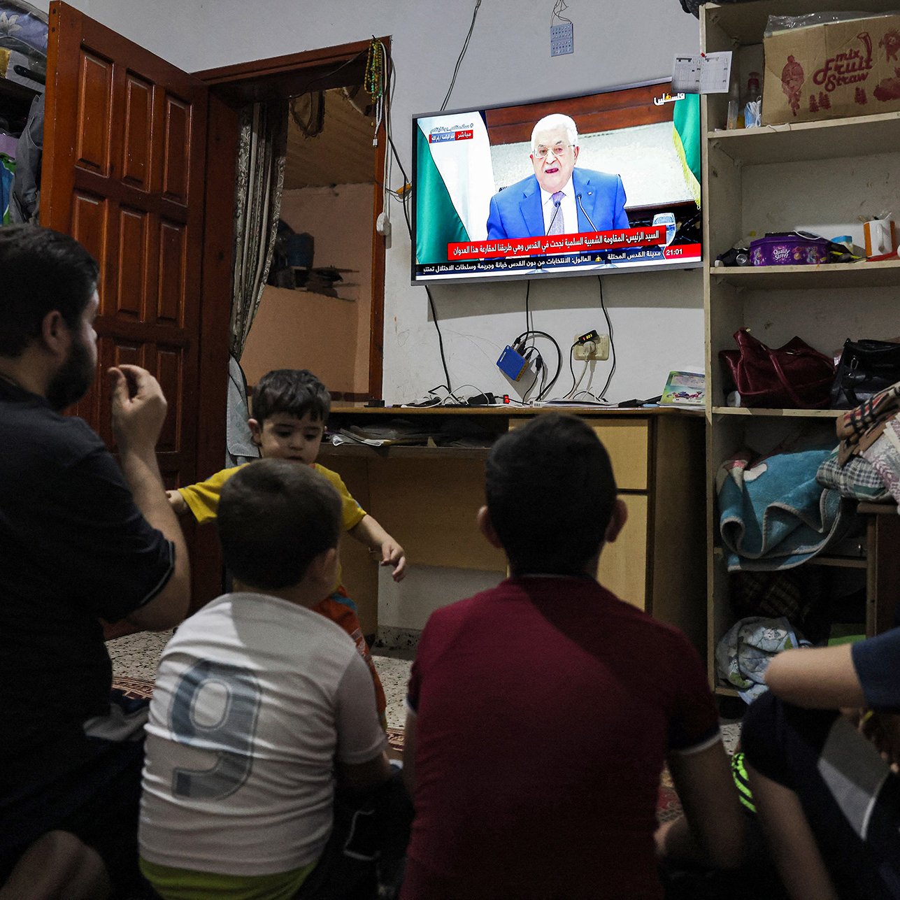 L'immagine mostra un gruppo di bambini seduti a terra, rivolti verso un televisore che trasmette un programma. Un adulto, probabilmente un genitore o un parente, è accanto a loro. La stanza sembra essere piccola e modesta, con mobili semplici e un'atmosfera informale. Sullo schermo si vede un uomo che parla, probabilmente un leader o un personaggio pubblico. I bambini appaiono curiosi e coinvolti nella visione.