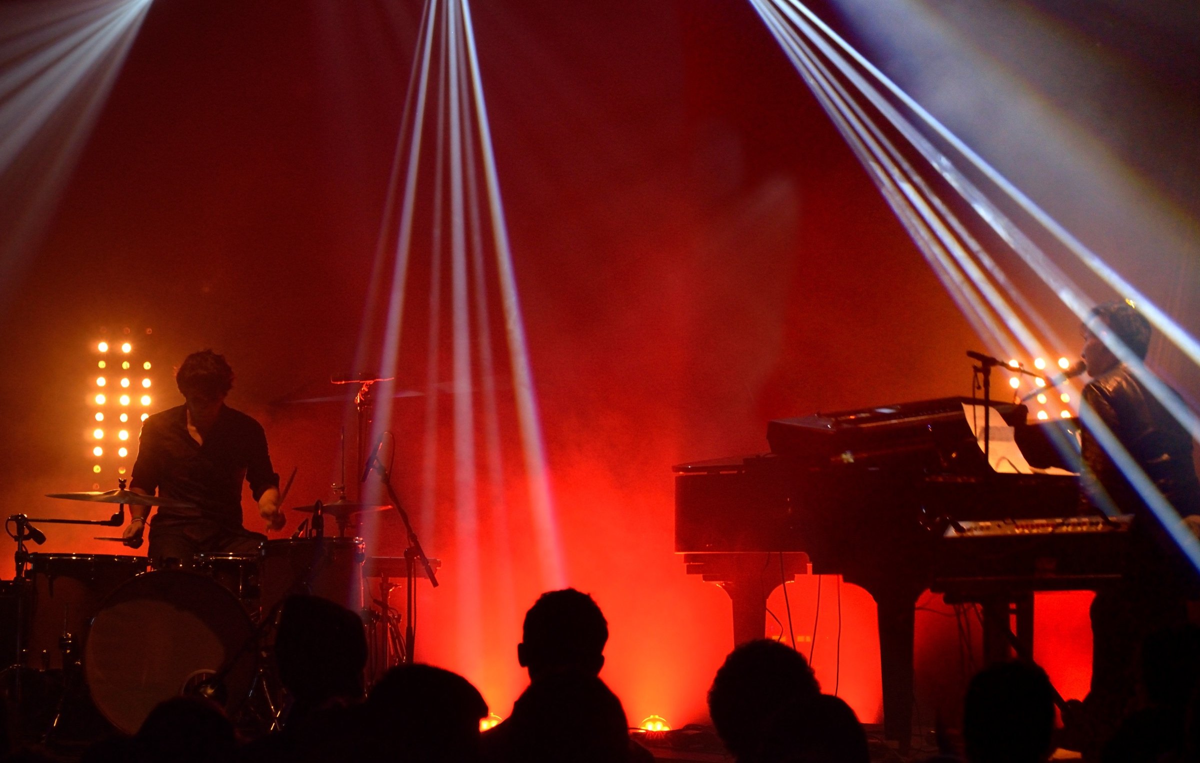 L'image montre une scène de concert avec deux musiciens. À gauche, un batteur joue de la batterie, tandis qu'à droite, un pianiste est assis devant un clavier. L'éclairage est dramatique, avec des faisceaux lumineux qui coupent à travers la fumée ambiante, créant une atmosphère dynamique. En arrière-plan, on devine une audience silhouette, profitant du spectacle. Les couleurs rouges et jaunes ajoutent à l'énergie de la performance.