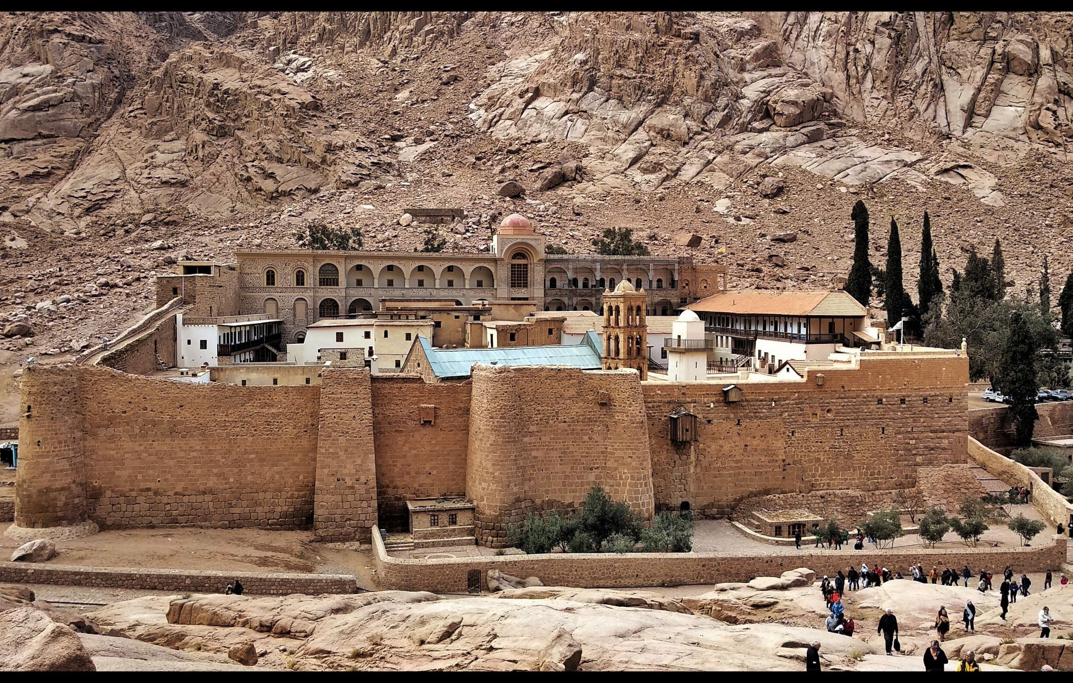 L'image montre un monastère situé dans une région montagneuse aride. Le bâtiment, de style architectural ancien, est construit en pierre et possède des murs épais. On peut voir une grande cour intérieure ainsi que des arbres qui ajoutent une touche de verdure au paysage. À l'arrière-plan, on distingue des formations rocheuses spectaculaires, soulignant l'isolement et la beauté naturelle des lieux. Des visiteurs se promènent autour du monastère, ce qui témoigne de son attrait touristique.