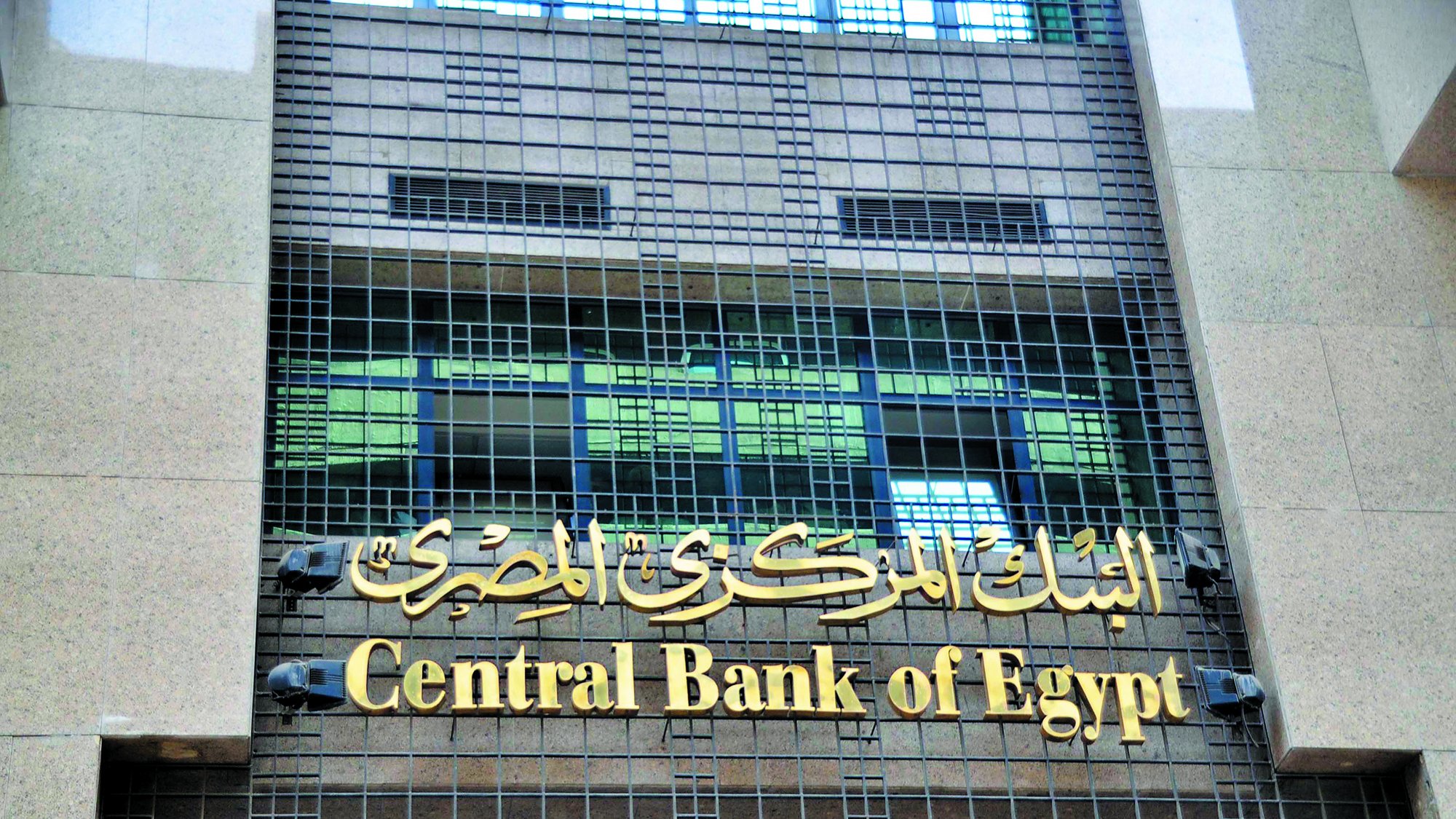 The image features the facade of a building that houses the Central Bank of Egypt. The building has a modern design with a prominent sign displaying "Central Bank of Egypt" in both English and Arabic. The Arabic text is situated above the English, and the overall appearance conveys a sense of official importance and stability. The building is constructed with stone materials, and its facade includes grid-like elements, suggesting a blend of contemporary and traditional architectural styles.