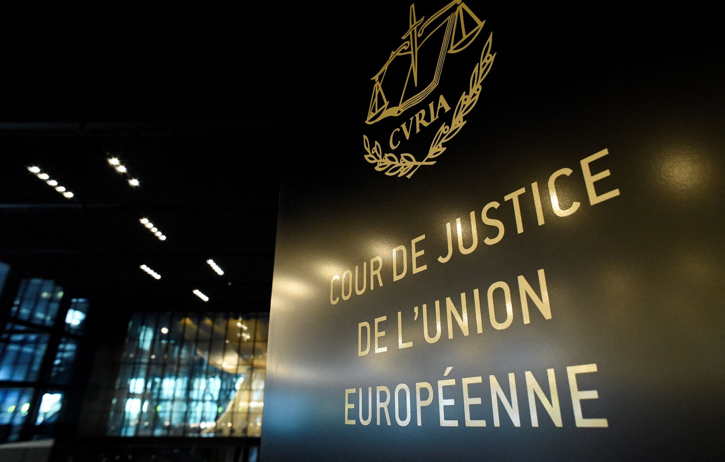L'image montre un panneau indiquant "COUR DE JUSTICE DE L'UNION EUROPÉENNE". Le panneau est stylisé avec des éléments graphiques, notamment une représentation de la balance de la justice. En arrière-plan, on peut voir une architecture moderne et intérieure, probablement d'un bâtiment officiel. L'éclairage est subtil, mettant en valeur le texte et le design du panneau.