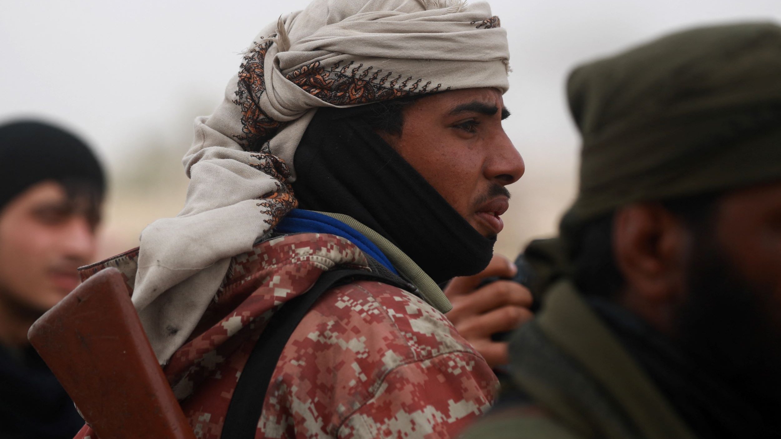 L'image montre un groupe d'individus, dont l'un est au premier plan. Il porte un couvre-chef traditionnel et une écharpe, ce qui lui donne un aspect distinctif. Son expression est sérieuse, et il semble concentré. Il tient également ce qui semble être une arme, indiquant un contexte potentiellement militaire ou de conflit. L'arrière-plan est flou, avec d'autres personnes visibles, suggérant une atmosphère de rassemblement ou d'affrontement.