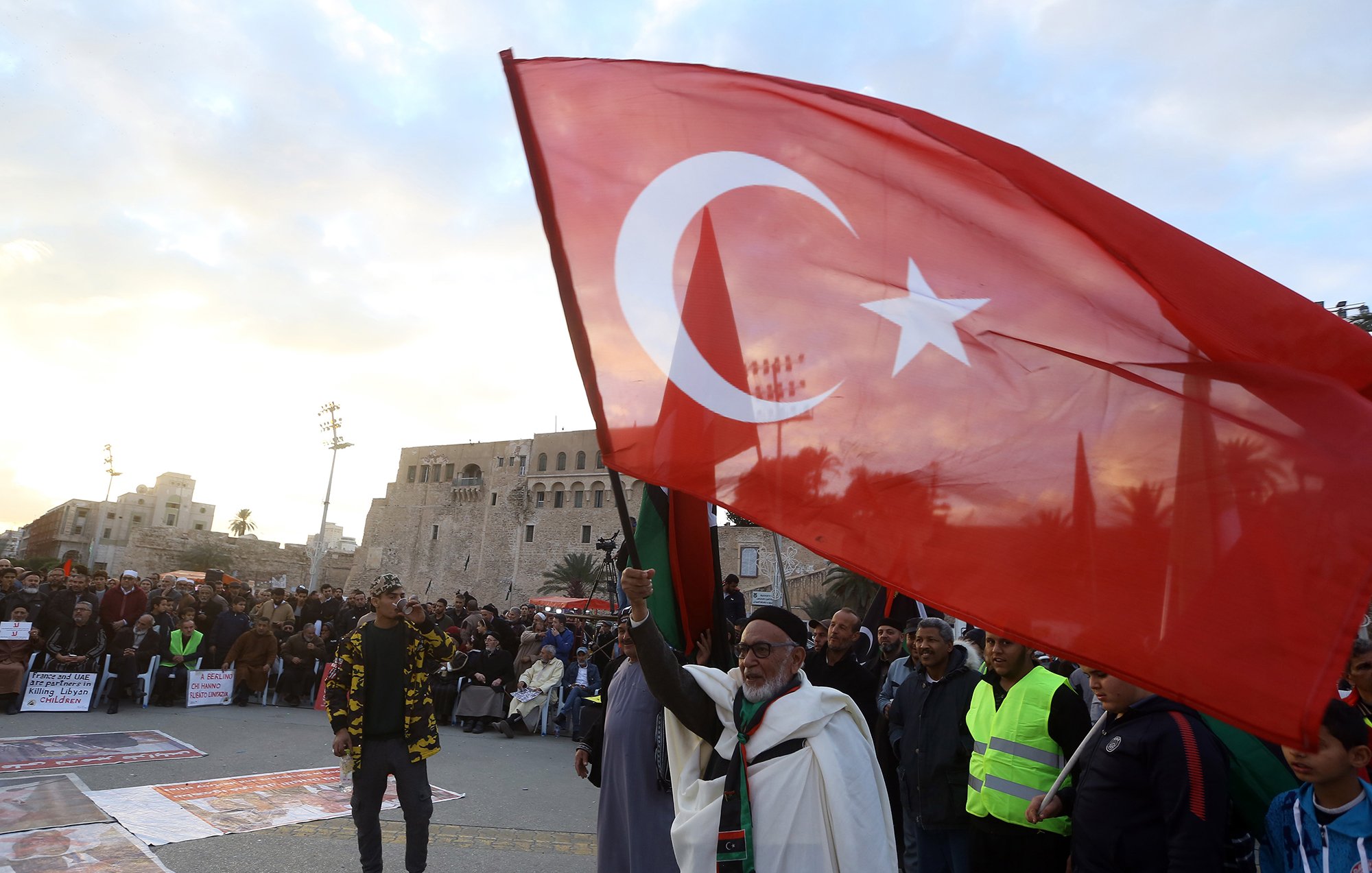 در این تصویر، جمعیتی در یک فضای باز مشاهده می‌شود که به نظر می‌رسد در حال برگزاری تجمعی هستند. یک مرد مسن با چهره‌ای شاد، پرچم بزرگ ترکیه را در دست دارد و به سوی دوربین اشاره می‌کند. در پس‌زمینه، ساختمان‌هایی قدیمی دیده می‌شود که نشان‌دهندهٔ تاریخ و فرهنگ خاصی هستند. مردم دیگری نیز در اطراف او به صورت جمعی ایستاده‌اند و تعدادی از آن‌ها لباس‌های زرد روشن پوشیده‌اند. آسمان ابری است و جلوه‌ای از نشاط و همبستگی در تصویر وجود دارد.