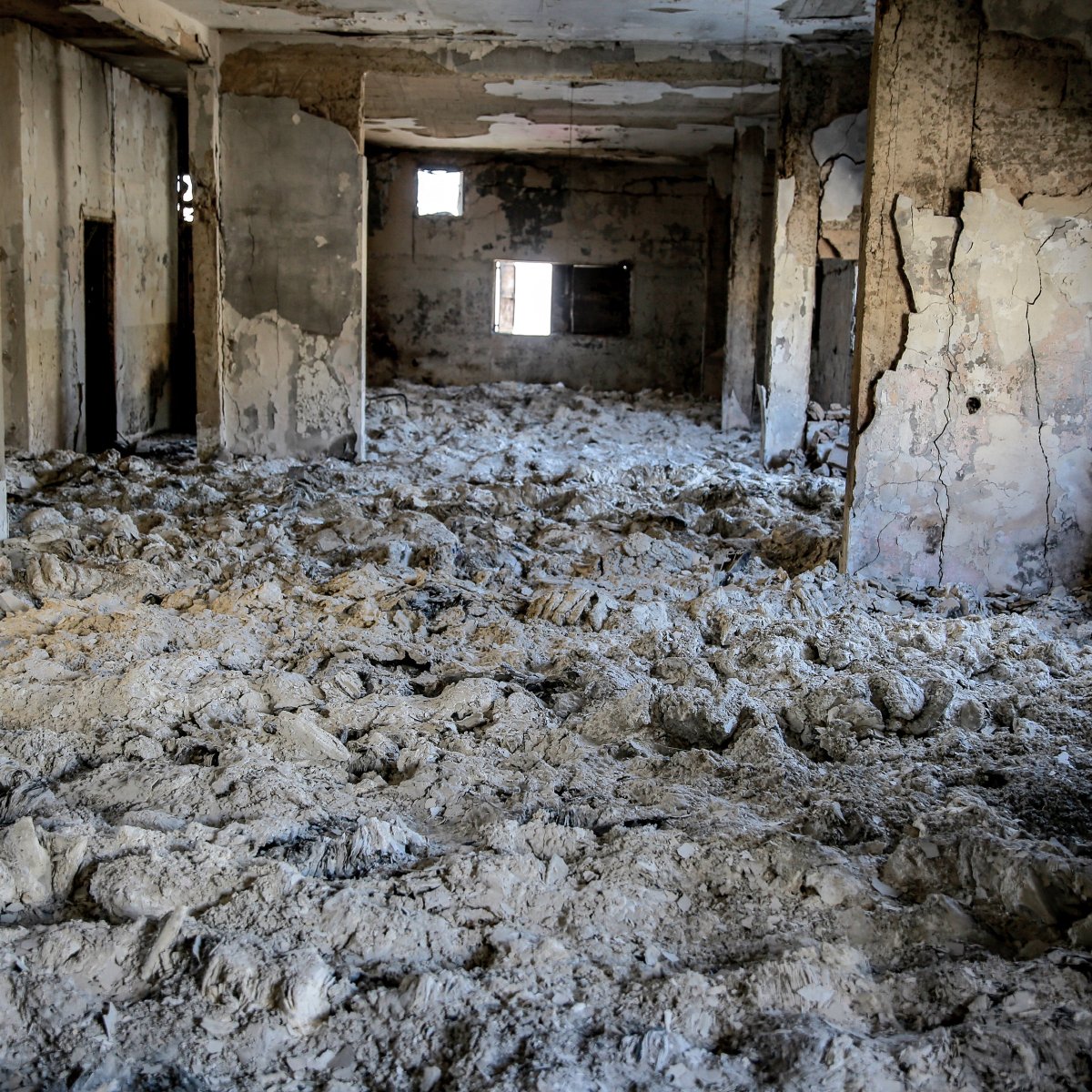 L'image montre l'intérieur d'un bâtiment en ruine, où les murs sont endommagés et décrépis. Le sol est recouvert d'un matériau blanc poussiéreux, probablement des débris ou des restes d'anciennes structures. Les fenêtres sont brisées ou manquantes, créant une atmosphère de désolation et d'abandon. La lumière pénètre à travers les ouvertures, mais l'espace parait sombre et sinistre.
