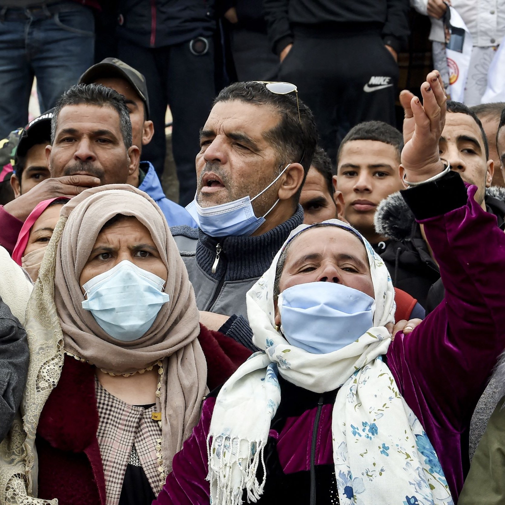 L'image montre une foule de personnes, principalement des hommes et des femmes, réunies pour exprimer leurs émotions. Certaines personnes portent des masques faciaux, et il y a des expressions de détermination et de protestation parmi les membres du groupe. Les vêtements sont variés, avec des accents de couleurs différentes. L'ambiance semble animée et engagée, suggérant une manifestation ou un rassemblement pour une cause.