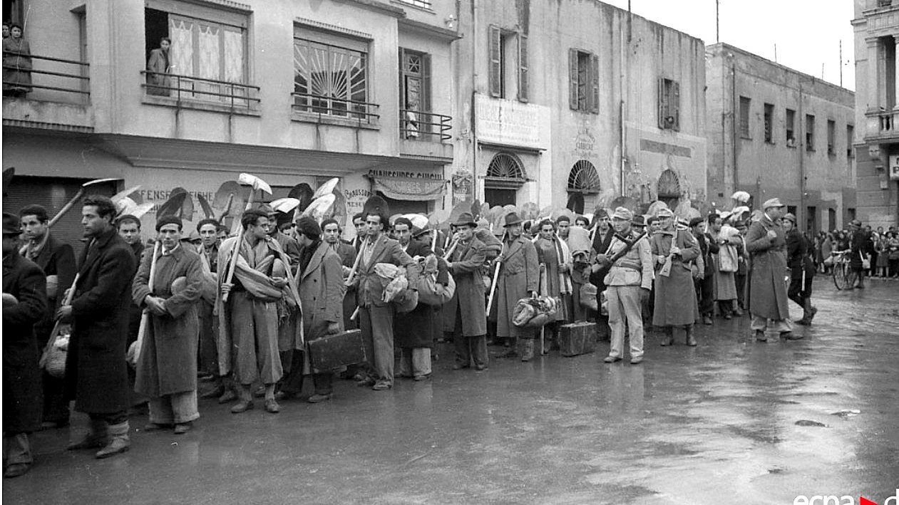 الصورة تظهر مجموعة من الأشخاص في صف طويل، يبدو أنهم ينتظرون شيئًا ما. يرتدون ملابس شتوية، ويحملون حقائب وأشياء أخرى. في الخلفية، يمكن رؤية مبانٍ تاريخية بشرفات ونوافذ. الجو يبدو هادئًا، ولكن المحيط يوحي بوجود حدث مهم أو تجمع. قد تعكس الصورة فترة زمنية معينة أو حدث اجتماعي.