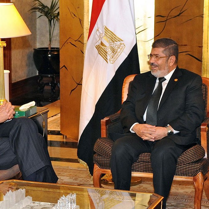 تظهر الصورة اجتماعًا بين شخصين يجلسان في غرفة مريحة. يجلس أحدهما على اليسار وهو يرتدي بدلة رسمية مع ربطة عنق ويبتسم، بينما يجلس الشخص الآخر على اليمين، كذلك يرتدي بدلة ويبدو سعيدًا. الخلفية تحتوي على علم مصر، مما يشير إلى أن الاجتماع قد يكون ذو طابع رسمي يتعلق بالشؤون السياسية أو العلاقات الدولية. الأجواء تبدو ودية وإيجابية.