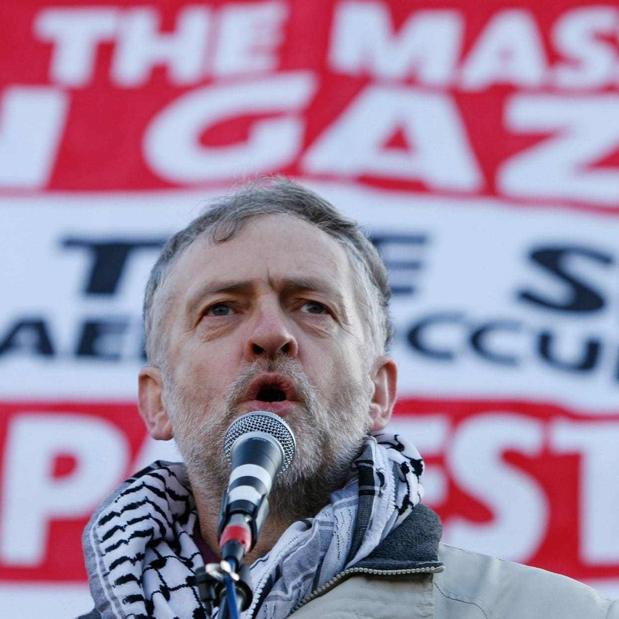 L'image montre un homme prenant la parole devant une grande banderole. Il porte un foulard traditionnel autour du cou et semble s'adresser à une foule. La banderole en arrière-plan contient des messages concernant la situation à Gaza, appelant à mettre fin aux massacres et à la occupation actuelle. L'expression de l'homme indique qu'il est engagé et passionné par le sujet qu'il aborde.