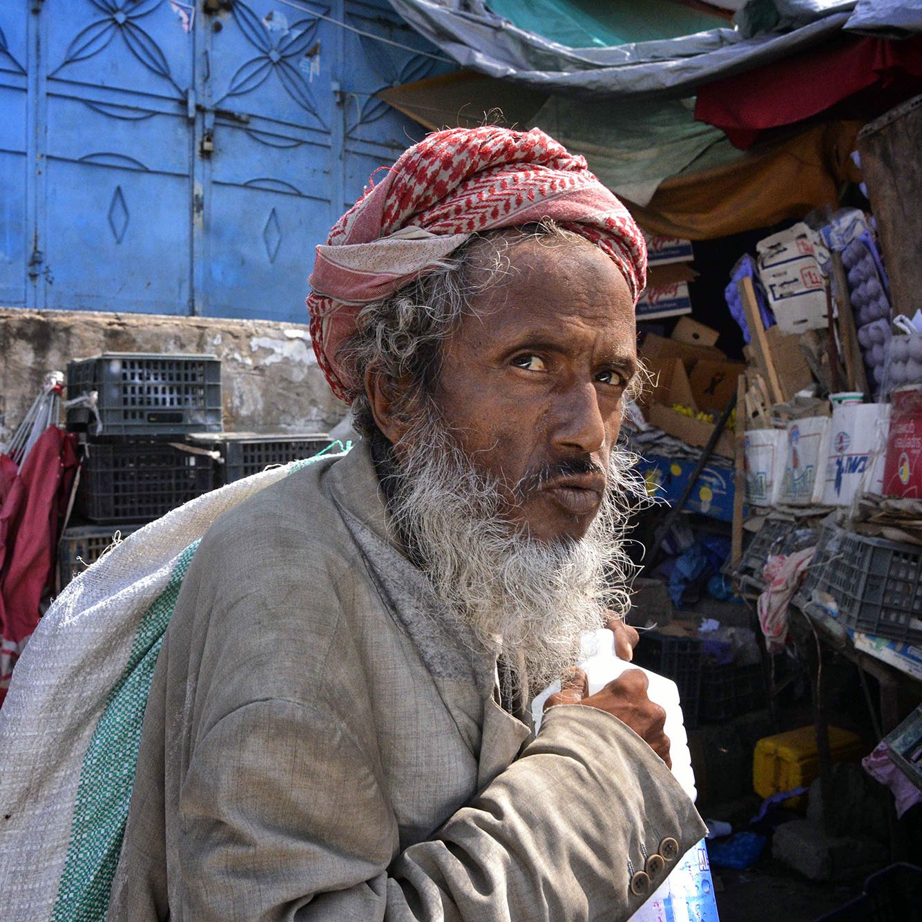 الصورة تُظهر رجلًا مسنًا، يبدو من ملامحه أنه يحمل تجاعيد تعبر عن تجارب الحياة. لديه لحية بيضاء وشعر تحت الغطاء الذي يضعه على رأسه. يرتدي ملابس بسيطة قديمة. تعكس الخلفية بساطتها وفوضاها، حيث توجد حاويات وخزائن وألوان متداخلة. تعبير وجهه يحمل مزيجًا من الحزن والتأمل، وكأنه يعيش في عالم خاص به.