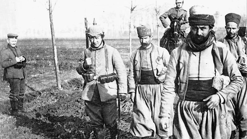 در این تصویر، گروهی از سربازان در حال راه رفتن در یک منطقه باز و مملو از گل و لای هستند. چند نفر از آن‌ها یونیفورم‌هایی به تن دارند که شامل شلوارهای گشاد و کلاه‌های خاص است. یکی از سربازان در حال حمل سلاح است و دیگری دستش را در جیبش قرار داده است. در پس‌زمینه، درختانی دیده می‌شوند که به تازگی کاشته شده‌اند. همچنین، یک نفر دیگر در گوشه تصویر ایستاده و به سمت گروه نگاه می‌کند. این تصویر به نظر می‌رسد که از یک دوره تاریخی خاص گرفته شده باشد و وضعیت نظامی را به نمایش می‌گذارد.