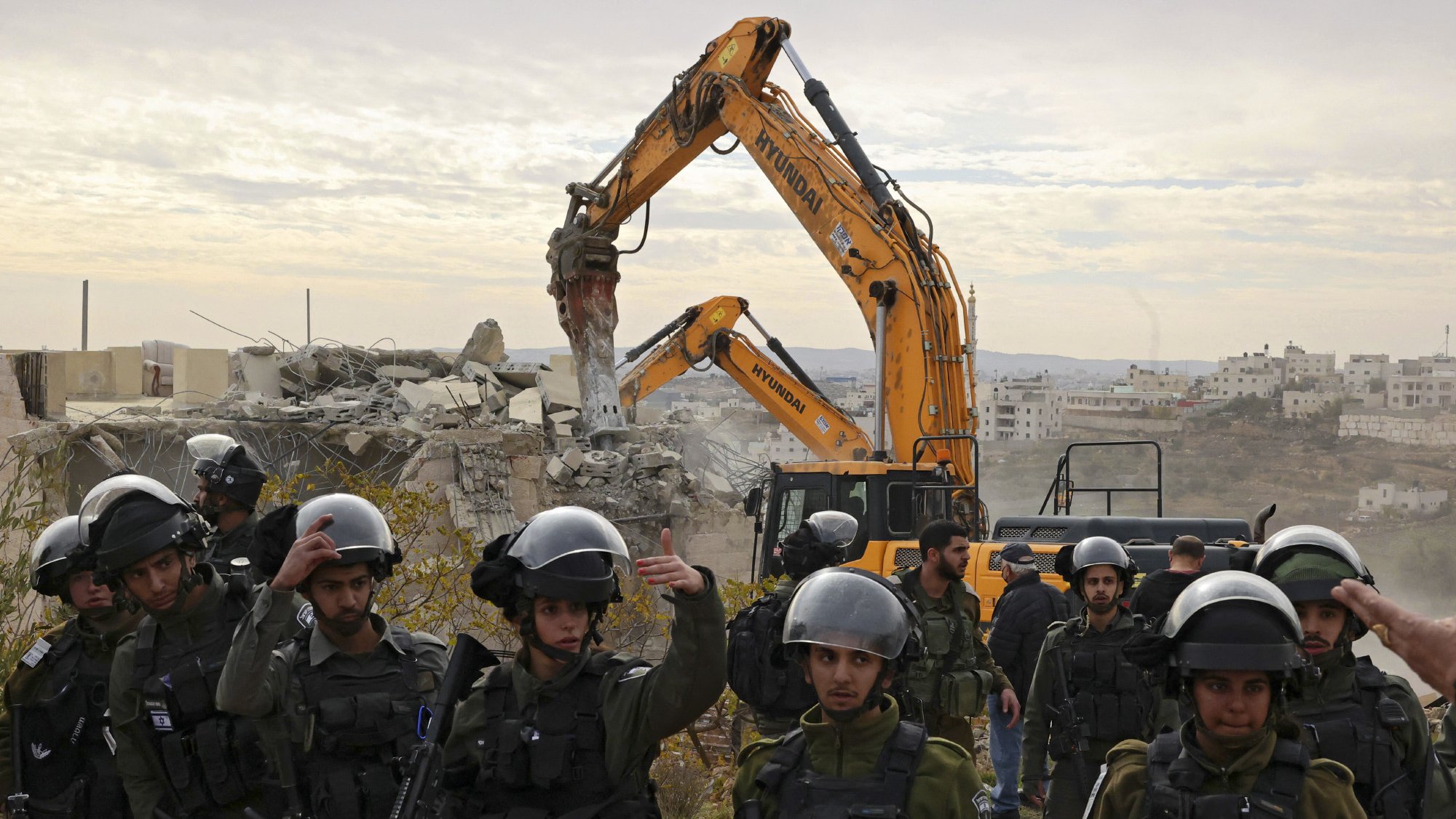 L'image montre une scène de destruction, où un bulldozer démolit un bâtiment en arrière-plan. Au premier plan, un groupe de soldats portant des casques et des équipements de protection surveille la situation. Ils semblent être en alerte, avec des armes en main. L'environnement est marqué par des décombres et un paysage urbain, suggérant un contexte de conflit. Le ciel est nuageux, ajoutant une atmosphère somber à la scène.