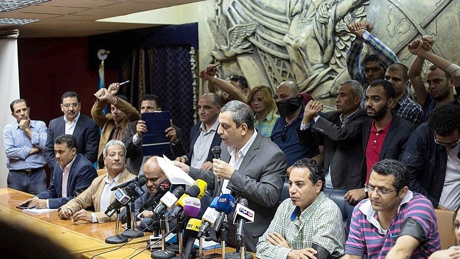 L'image montre une conférence de presse ou une réunion. Un homme se tient au centre, parlant devant un groupe de journalistes, avec plusieurs microphones devant lui. Derrière lui, des personnes semblent exprimer leur soutien ou leur désaccord, levant les poings. L'arrière-plan présente une sculpture en relief qui ajoute de la profondeur à la scène. L'atmosphère semble tendue et engagée.