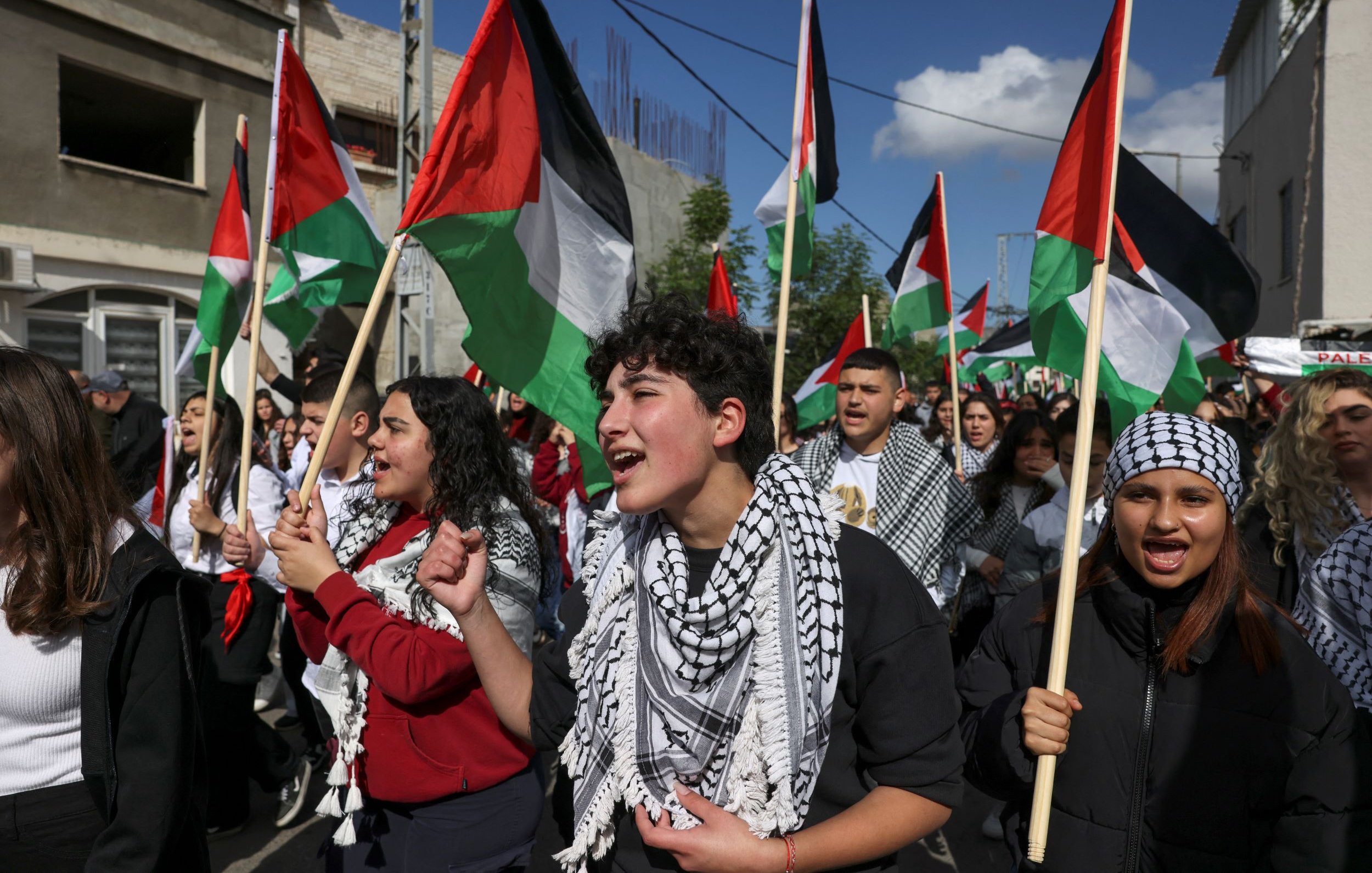 L'image montre une foule de manifestants, principalement des jeunes, participant à une marche ou une manifestation. Ils portent des drapeaux palestiniens aux couleurs rouge, noir, blanc et vert, et certains portent des keffiehs, des écharpes traditionnelles palestiniennes, souvent à motifs noirs et blancs. Les manifestants semblent passionnés et chantent, exprimant leur soutien à une cause. En arrière-plan, on aperçoit des bâtiments et un ciel partiellement nuageux.