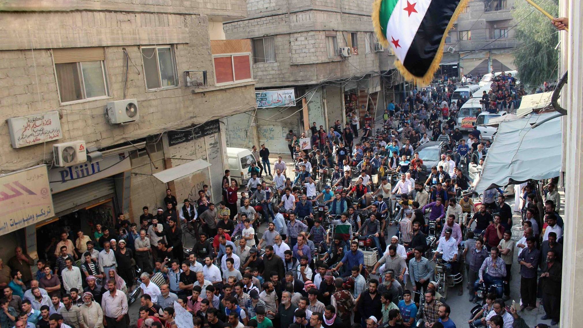 L'image montre une grande foule rassemblée dans une rue, probablement dans une ville syrienne. Les gens semblent se réunir pour une manifestation ou un événement public, avec des pancartes et un drapeau aux couleurs vives, typiquement associé à des mouvements de protestation. Les bâtiments environnants affichent des signes de vie urbaine, et plusieurs véhicules sont stationnés dans la rue. L'atmosphère semble intensément animée, avec une forte concentration de personnes dans l'espace.
