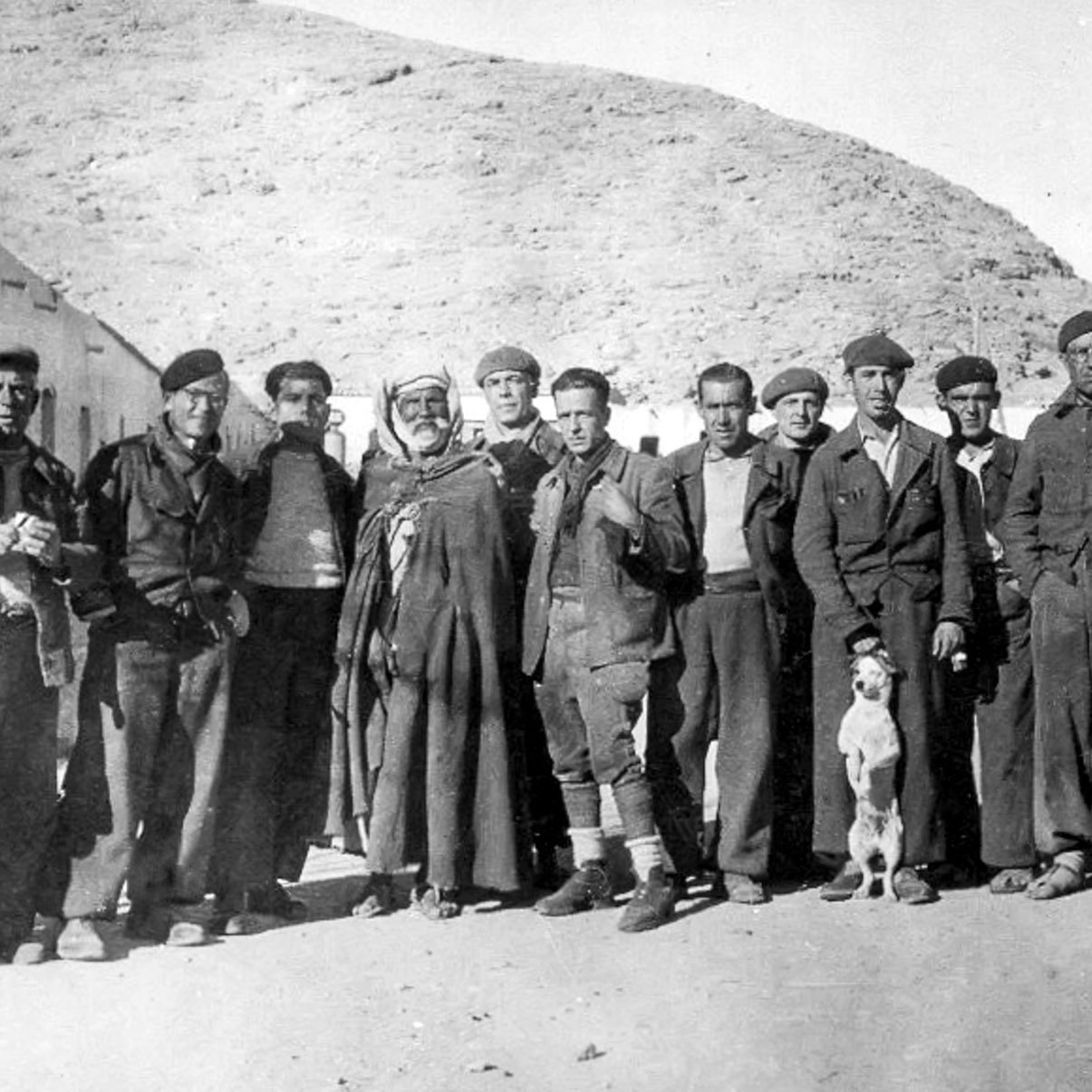 الصورة تظهر مجموعة من الأشخاص الذين يقفون معاً في مكان مفتوح، يبدو أنهم مجموعة تاريخية أو ربما فريق عمل. يرتدي معظمهم ملابس خاصة، مثل الزي العسكري أو ملابس العمل، وبعضهم يحمل أكواباً أو أدوات. في وسط المجموعة يوجد رجل يرتدي غطاء رأس تقليدي. هناك أيضاً كلب صغير يقف بين الأشخاص. تبدو الخلفية جبلية، مما يشير إلى أنهم في منطقة ريفية أو جبلية. تعكس الصورة أجواء من زمن بعيد وتحمل طابعاً تاريخياً.