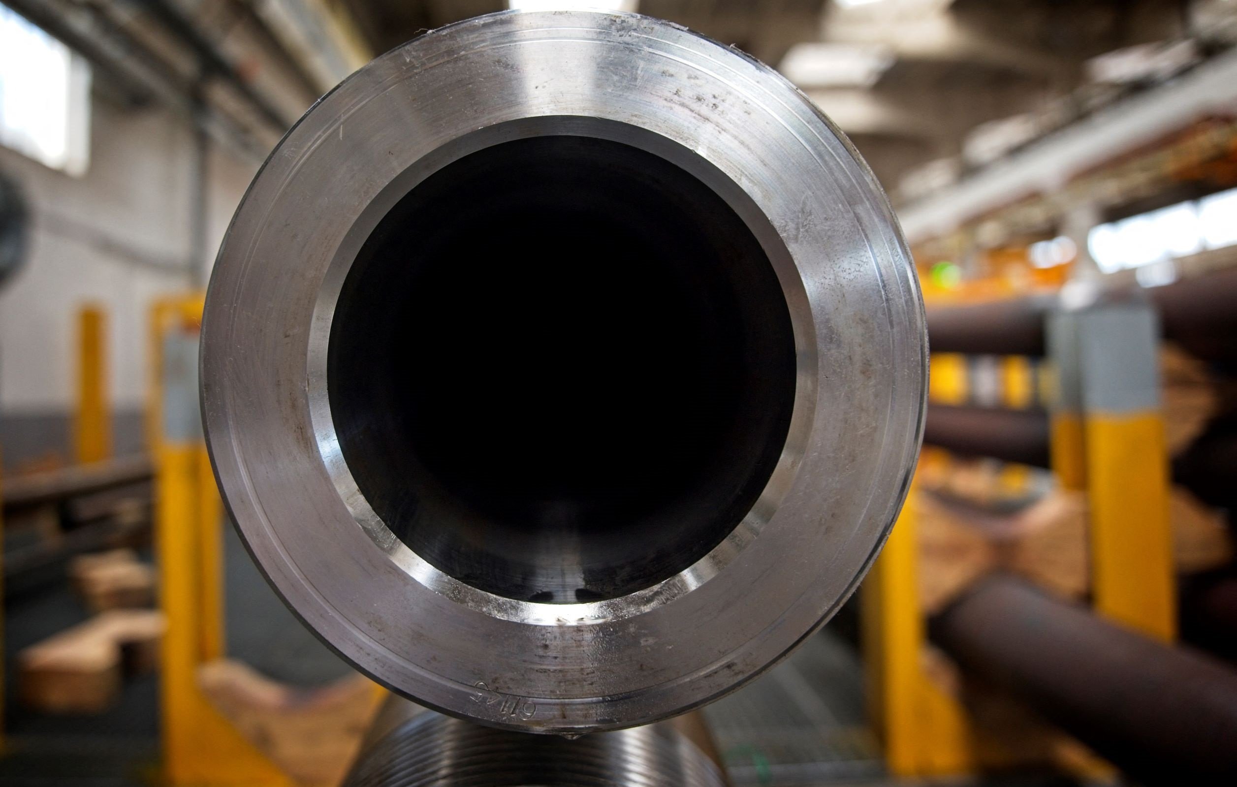 The image features a close-up view of a large metallic pipe, focusing on the open end. The interior of the pipe appears dark and hollow, creating a stark contrast with the shiny, reflective surface around the opening. In the background, there are other pipes and industrial elements, suggesting a manufacturing or storage environment. The lighting implies a spacious industrial setting, with hints of machinery and equipment visible farther back.