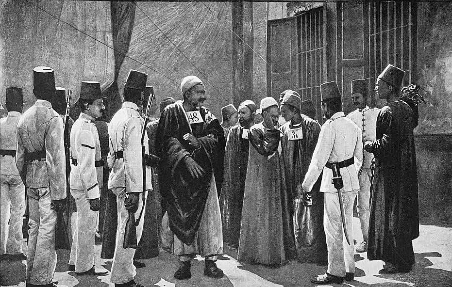 L'image présente un groupe de personnes en tenue traditionnelle. Au centre, un homme en robe longue semble diriger la scène, tandis que d'autres, habillés en uniformes ou en costumes distinctifs, l'entourent. Certains des individus portent des chapeaux spécifiques, comme des fez. Ils semblent engagés dans une conversation ou une activité, suggérant une interaction communautaire ou une réunion formelle. L'arrière-plan indique un environnement urbain, peut-être une scène historique. La composition dégage une atmosphère d'autorité et de respect.