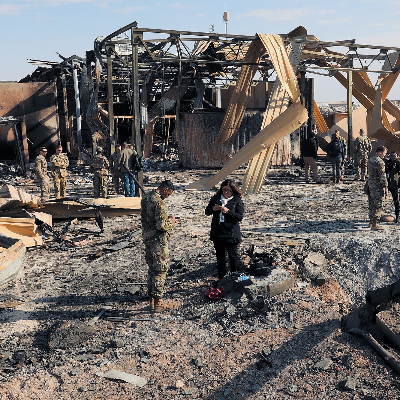 L'image montre une scène de destruction, probablement après un bombardement ou un incendie. On peut voir des débris et des structures en ruines, avec des panneaux de bois fissurés. Plusieurs militaires et civils se trouvent sur le site, certains en train de prendre des notes ou d'observer les dégâts. L'atmosphère semble grave, soulignant les conséquences d'un conflit. Le ciel est clair, contrastant avec le désastre au sol.