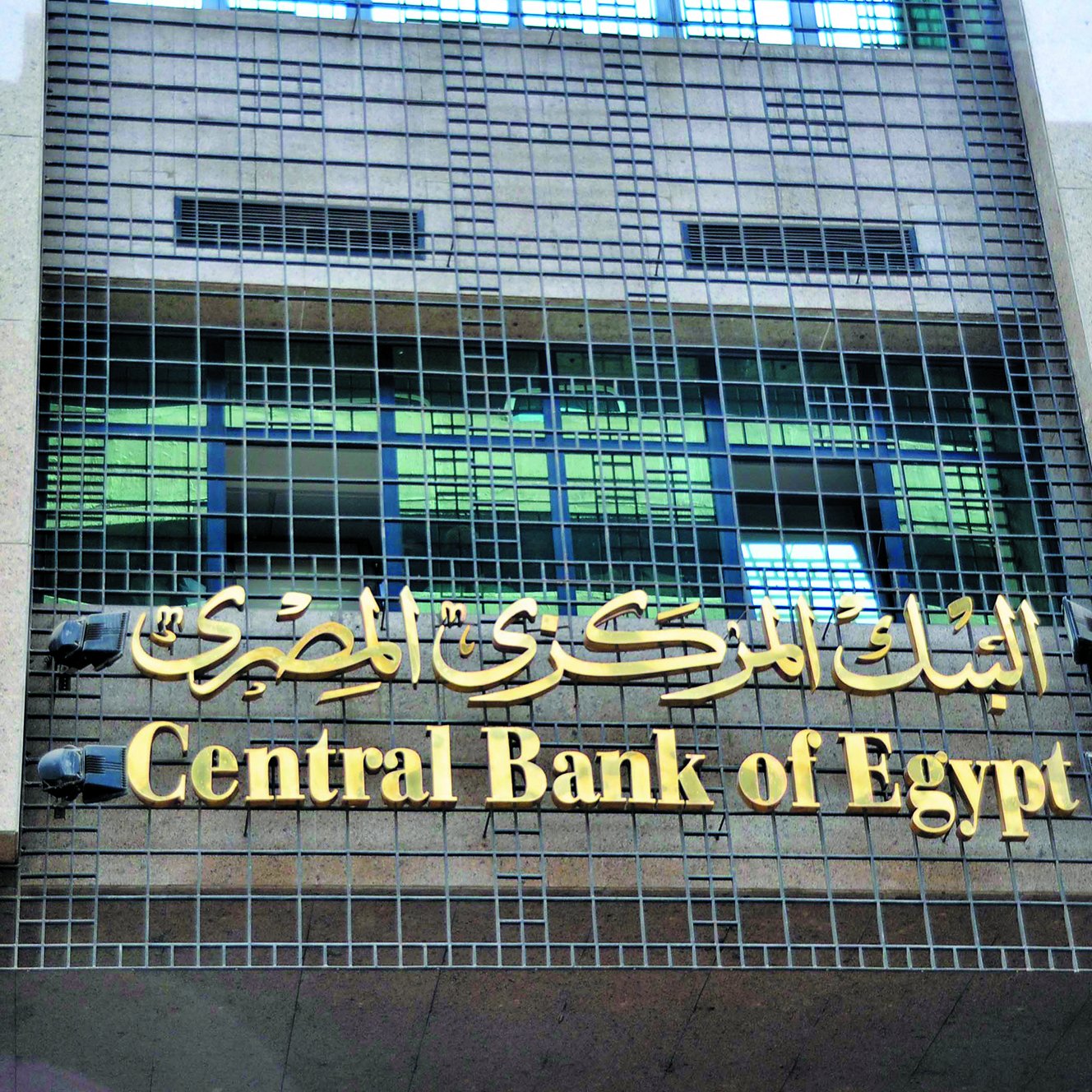 الصورة تظهر واجهة البنك المركزي المصري. يتكون التصميم من لافتة تحمل اسم البنك مكتوبًا باللغة العربية "البنك المركزي المصري" وباللغة الإنجليزية "Central Bank of Egypt". المبنى يبدو حديثًا ويستخدم الزجاج والجرانيت في واجهته، مما يعطيه مظهرًا أنيقًا ورسميًا.