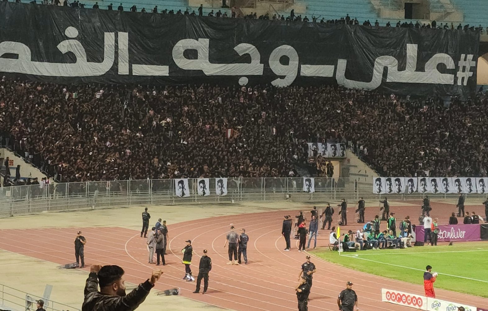 Stade plein de supporters en noir avec une grande banderole en arabe. Énergie et passion.