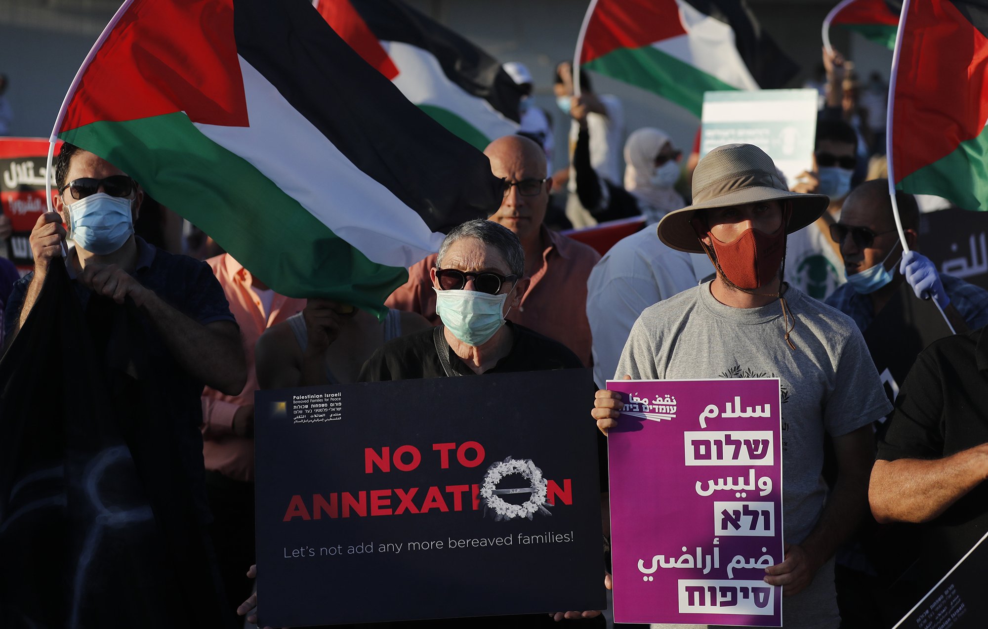 L'image montre une manifestation avec des participants portant des masques. Plusieurs personnes tiennent des drapeaux palestiniens. Des pancartes sont visibles, certaines affichant des messages tels que "NO TO ANNEXATION" (Non à l'annexion) et d'autres slogans en arabe et en hébreu. L'ambiance semble être celle d'une mobilisation pour une cause sociale ou politique, avec un engagement visible des manifestants.