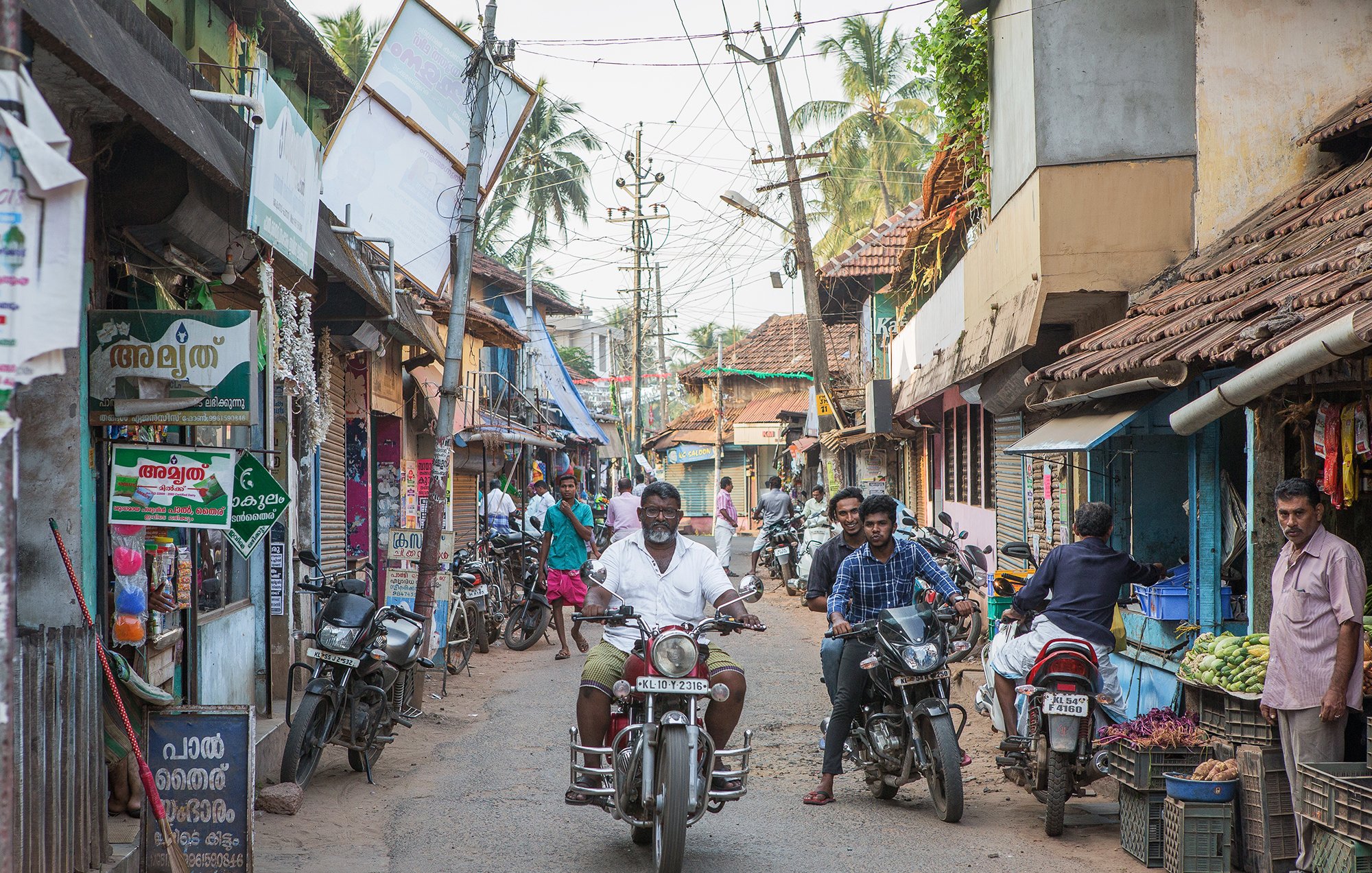 تظهر الصورة مشهداً من شارع ضيق في منطقة ريفية أو شبه حضرية. يتواجد في الصورة عدد من الأشخاص يسيرون ويتحدثون، بينما يقود رجل دراجة نارية في منتصف الشارع. المباني على الجانبين بسيطة ومكونة من طوابق قليلة، مع أسطح مغطاة بالبلاط. يمكن رؤية لافتات ملونة على المحلات التجارية التي تبيع مختلف السلع، وهناك أشجار تتواجد في الخلفية مما يعطي انطباعاً بحياة محلية نابضة. الجو يبدو هادئاً ومعتاداً على النشاط اليومي للسكان.