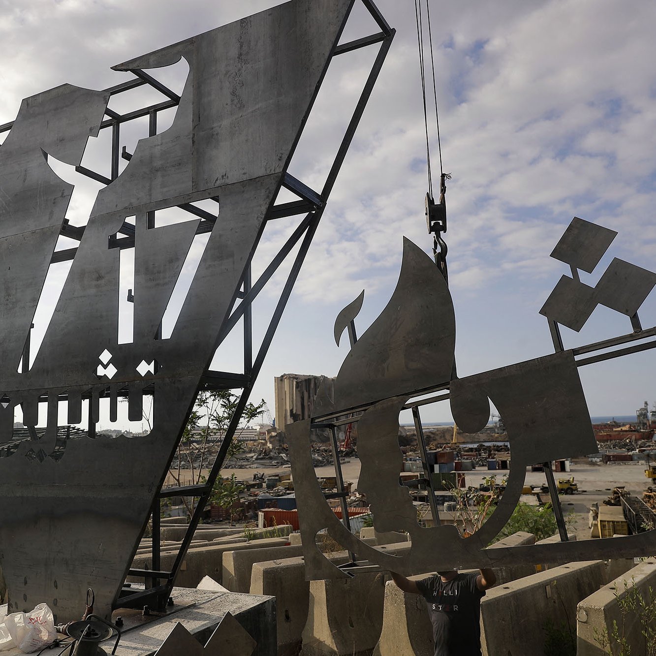 L'image montre une œuvre d'art en acier, probablement une sculpture moderne, qui est en cours d'installation ou de fabrication. La sculpture présente des formes géométriques et abstraites, avec des motifs qui semblent représenter des éléments architecturaux ou des symboles culturels. En arrière-plan, on aperçoit un chantier ou un port, avec des équipements de construction et des bateaux. Le ciel est partiellement nuageux, ajoutant une ambiance dynamique à la scène.