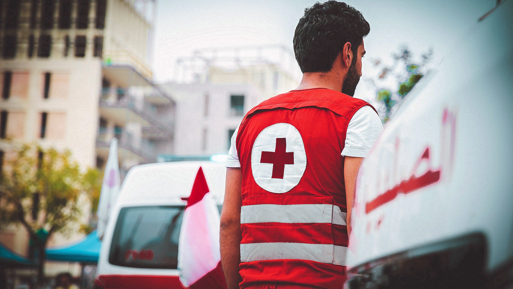 الصورة تظهر شابًا يرتدي زيًا خاصًا بالهلال الأحمر، والذي يتكون من سترة حمراء مزينة بعلامة الصليب الأحمر. يبدو الشاب من الخلف، وهو يقف بالقرب من سيارات إسعاف تحمل شعار الهلال الأحمر. تظهر في الخلفية مباني ومرافق عامة، مما يشير إلى وجود أنشطة أو أحداث إنسانية في المنطقة. تعكس الصورة الجهد الإنساني ورغبة المساعدة في الحالات الطارئة.