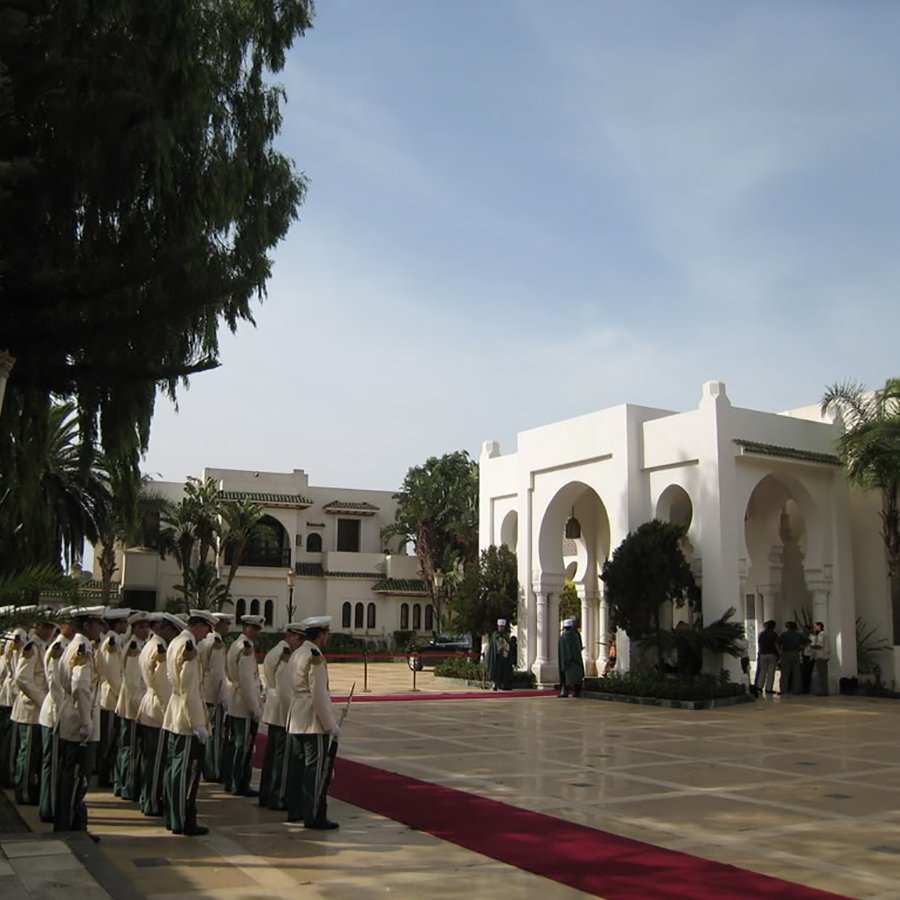 L'image montre une scène extérieure d'un bâtiment élégant, probablement un palais ou un hôtel de luxe. On peut voir une allée centrale avec un tapis rouge, entourée de verdure et de palmiers. À gauche, un groupe de personnes en uniformes formés, représentant une sorte de garde ou de cérémonie. En arrière-plan, on aperçoit un bâtiment majestueux avec des arches et des colonnes, suggérant une architecture raffinée. Le ciel est clair, ce qui ajoute à l'atmosphère solennelle de la scène.