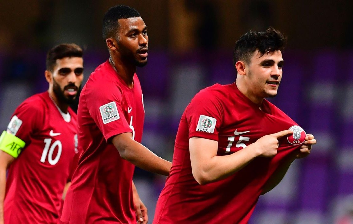 L'image montre des joueurs de football célébrant un moment de victoire ou d'accomplissement sur le terrain. Ils portent des maillots rouges avec des logos de l'équipe, et l'un d'eux semble montrer un symbole ou une médaille en soulevant son maillot. Les autres joueurs réagissent joyeusement derrière lui, montrant l'esprit d'équipe et la camaraderie. L'arrière-plan fait référence à un stade, créant une atmosphère d'événement sportif.