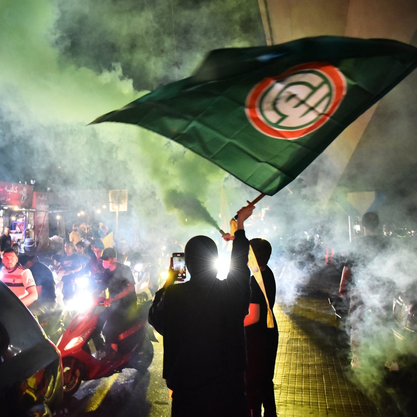 مردم در خیابان با پرچم‌های سبز و زرد، در میان دود و آتش‌بازی، جشن و اعتراض می‌کنند.
