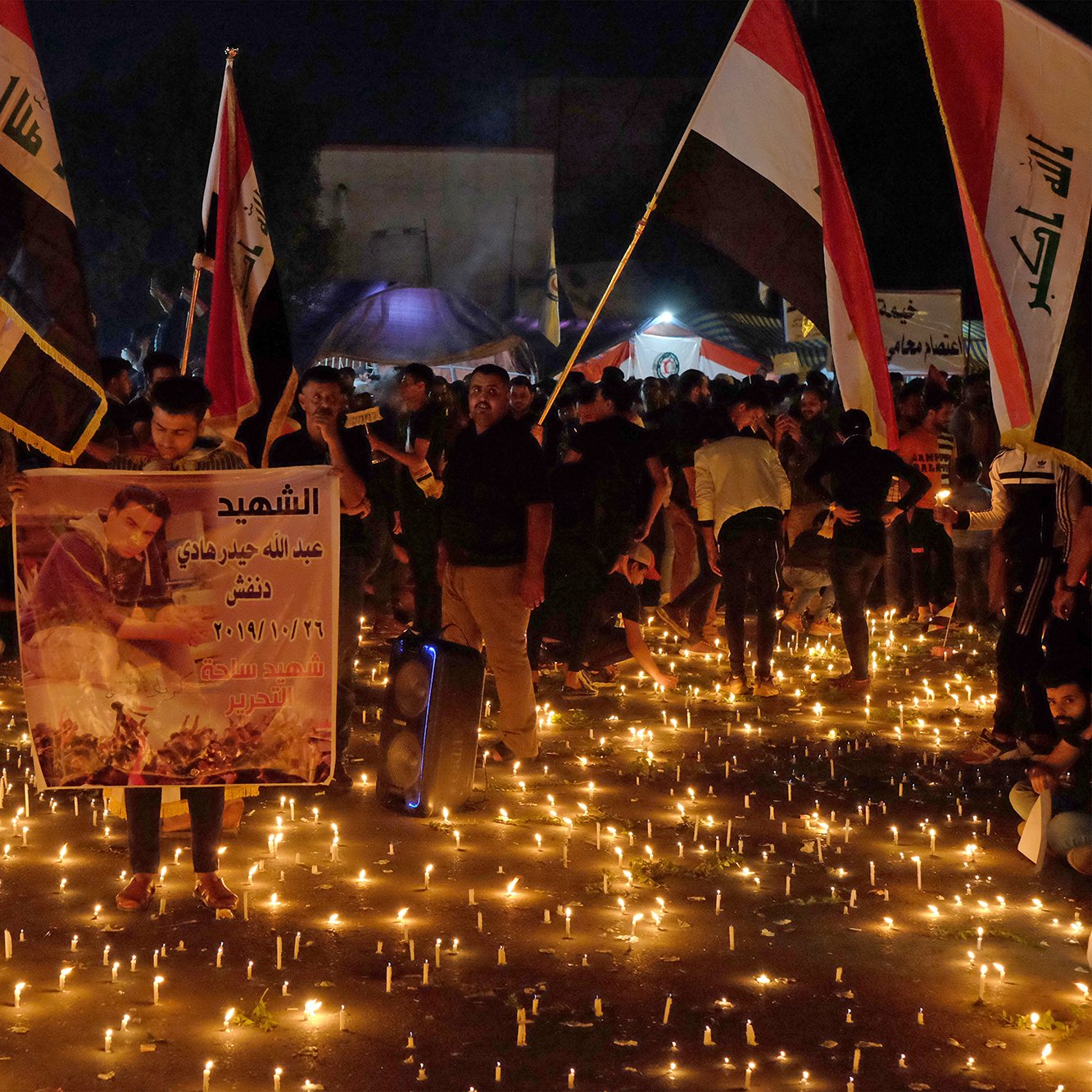 توضح الصورة تجمعًا حاشدًا للناس في مكان مفتوح خلال الليل، حيث يضيء المكان بكثرة الشموع الموزعة على الأرض. يحمل المشاركون لافتات وصورًا، وبعضهم يرفع أعلامًا تمثل بلدانهم. الجو يبدو مهيبًا، حيث يعبر الناس عن حزنهم أو إحياء ذكرى معينة، مما يعكس شعورًا بالوحدة والتضامن في هذه اللحظة.