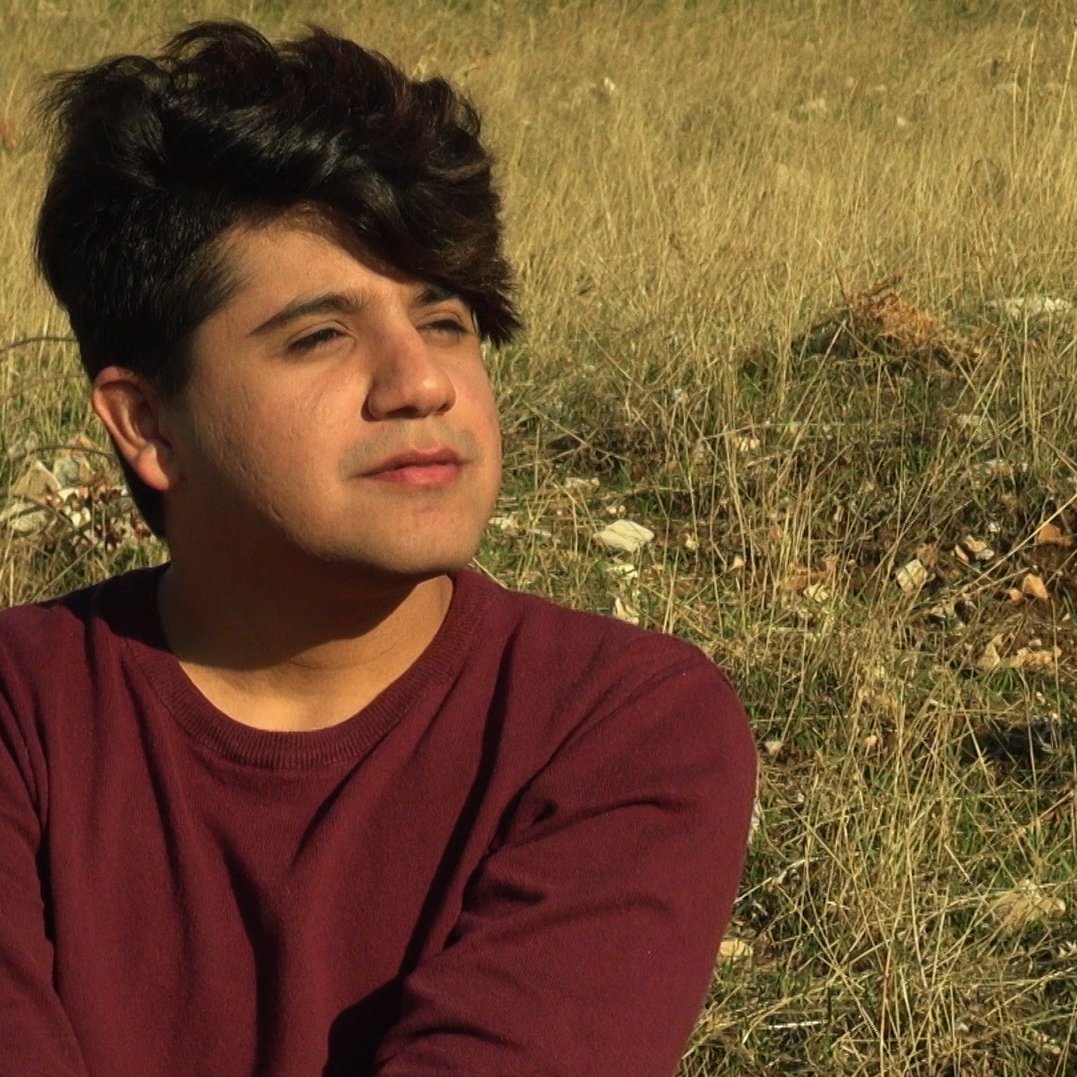 L'image montre une personne assise dans un champ de herbes hautes. La personne a les cheveux foncés et bouclés, et elle semble pensive, regardant au loin. Elle porte un pull de couleur bordeaux. L'environnement est naturel, avec une lumière douce, ce qui crée une atmosphère tranquille.