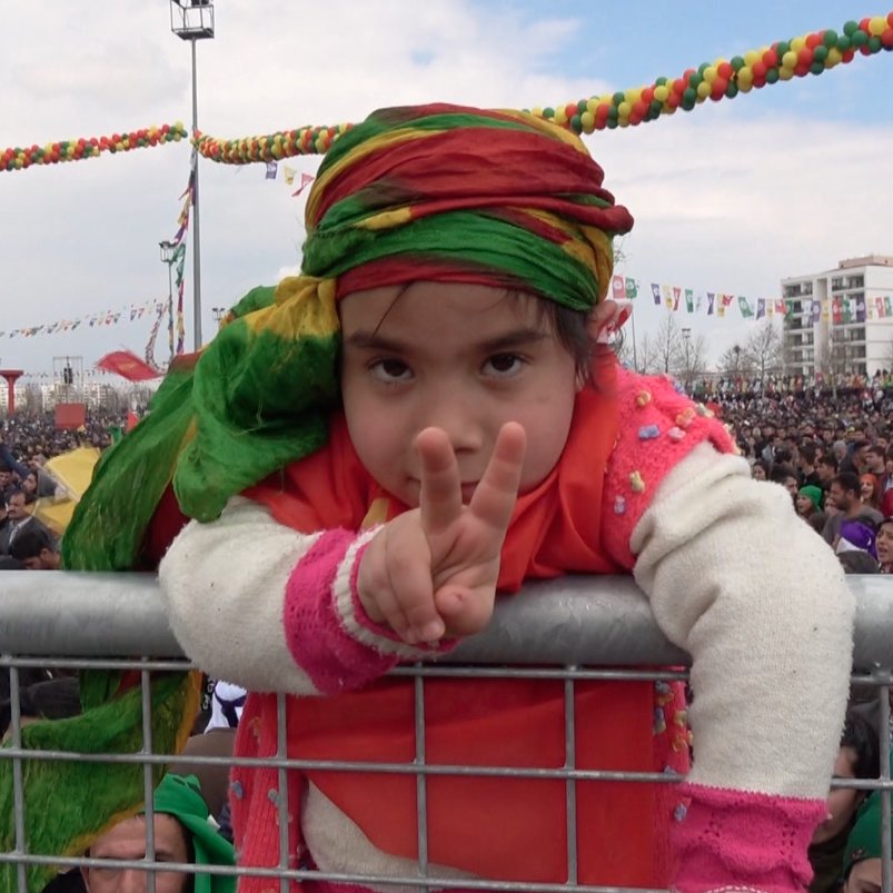 L'image montre une jeune fille participant à un grand rassemblement. Elle est vêtue de vêtements colorés, portant un foulard aux teintes vives. La fillette fait un signe de paix avec ses doigts, pendant qu'elle se penche sur une barrière. En arrière-plan, une grande foule est présente, témoignant d'un événement festif ou d'une manifestation populaire, avec des décorations colorées suspendues au-dessus. L'ambiance semble joyeuse et engagée.