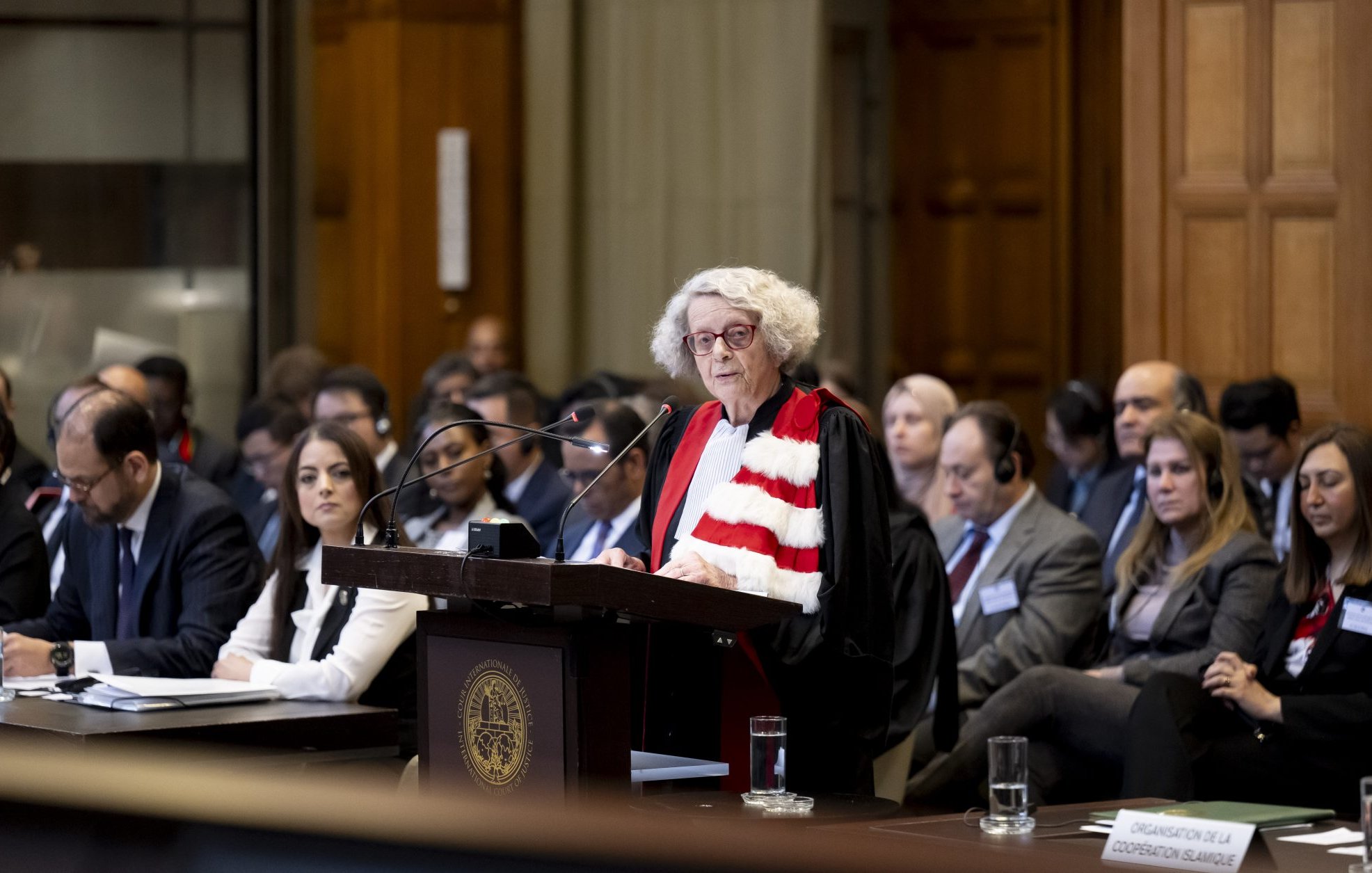 L'image montre une scène dans une salle d'audience, probablement dans un tribunal. Une femme, vêtue d'une robe magistrale et d'une écharpe, se tient derrière un pupitre en train de prendre la parole. Plusieurs personnes sont assises à des tables devant elle, écoutant attentivement. L'environnement semble formel, avec des éléments architecturaux en bois et des fenêtres qui apportent de la lumière naturelle. L'atmosphère est solennelle et concentrée, indicative d'une procédure judiciaire importante.