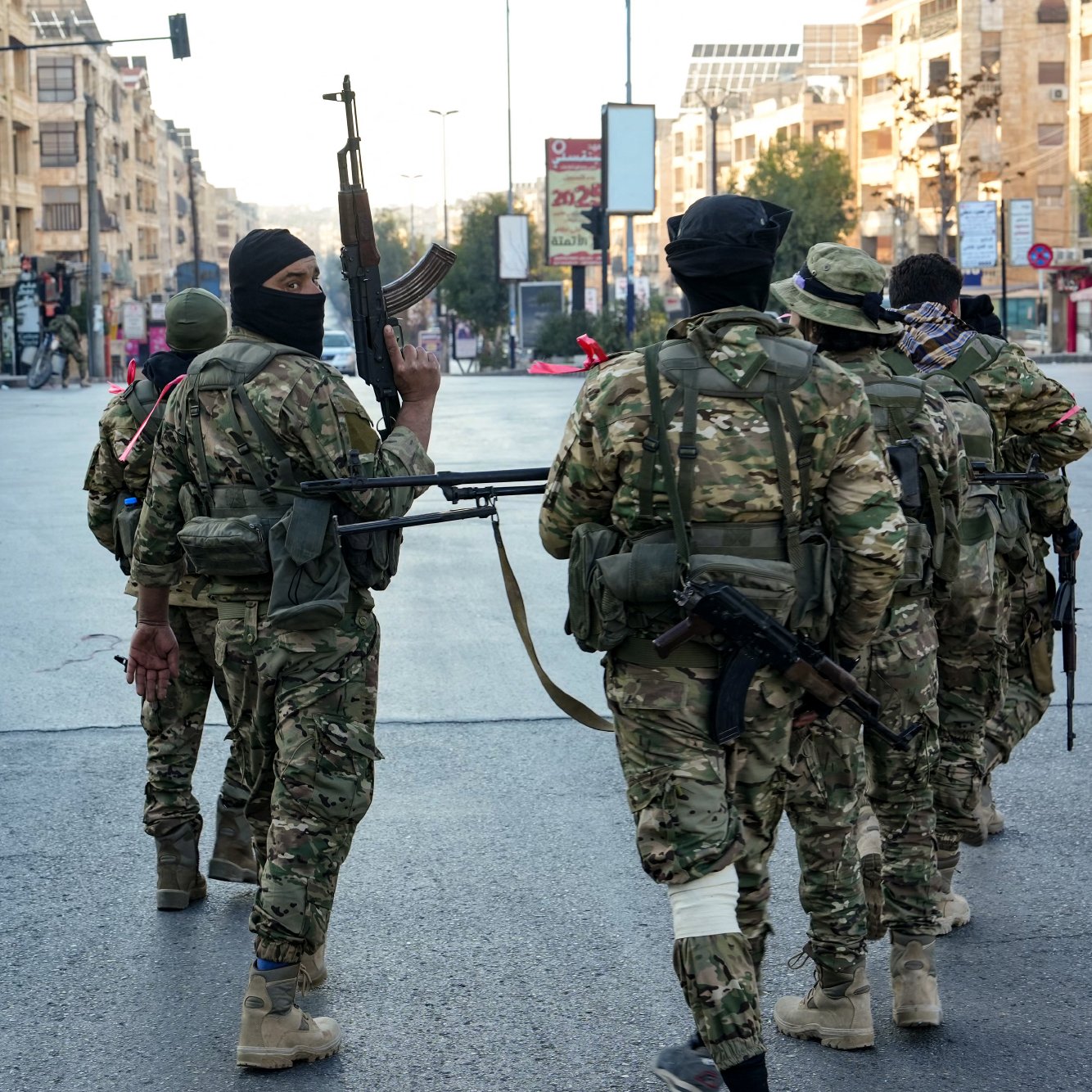 L'immagine mostra un gruppo di soldati in abbigliamento militare che camminano in una strada urbana. I soldati sono armati e indossano maschere che coprono parte del volto. Sullo sfondo si possono vedere edifici e un ambiente urbano, con luci e segnali stradali visibili. L'atmosfera è quella di un contesto di sicurezza o di operazioni militari.