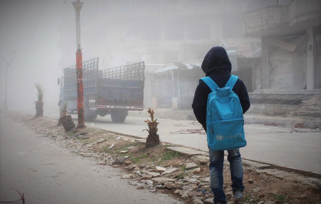L'image montre un jeune garçon de dos, portant un grand sac à dos bleu. Il se tient sur une rue où la visibilité est réduite en raison d'un épais brouillard. En arrière-plan, on peut apercevoir des bâtiments partiellement détruits et un camion stationné. Le sol est jonché de débris et de petites plantes. L'atmosphère est sombre et mélancolique, évoquant une scène de désolation.