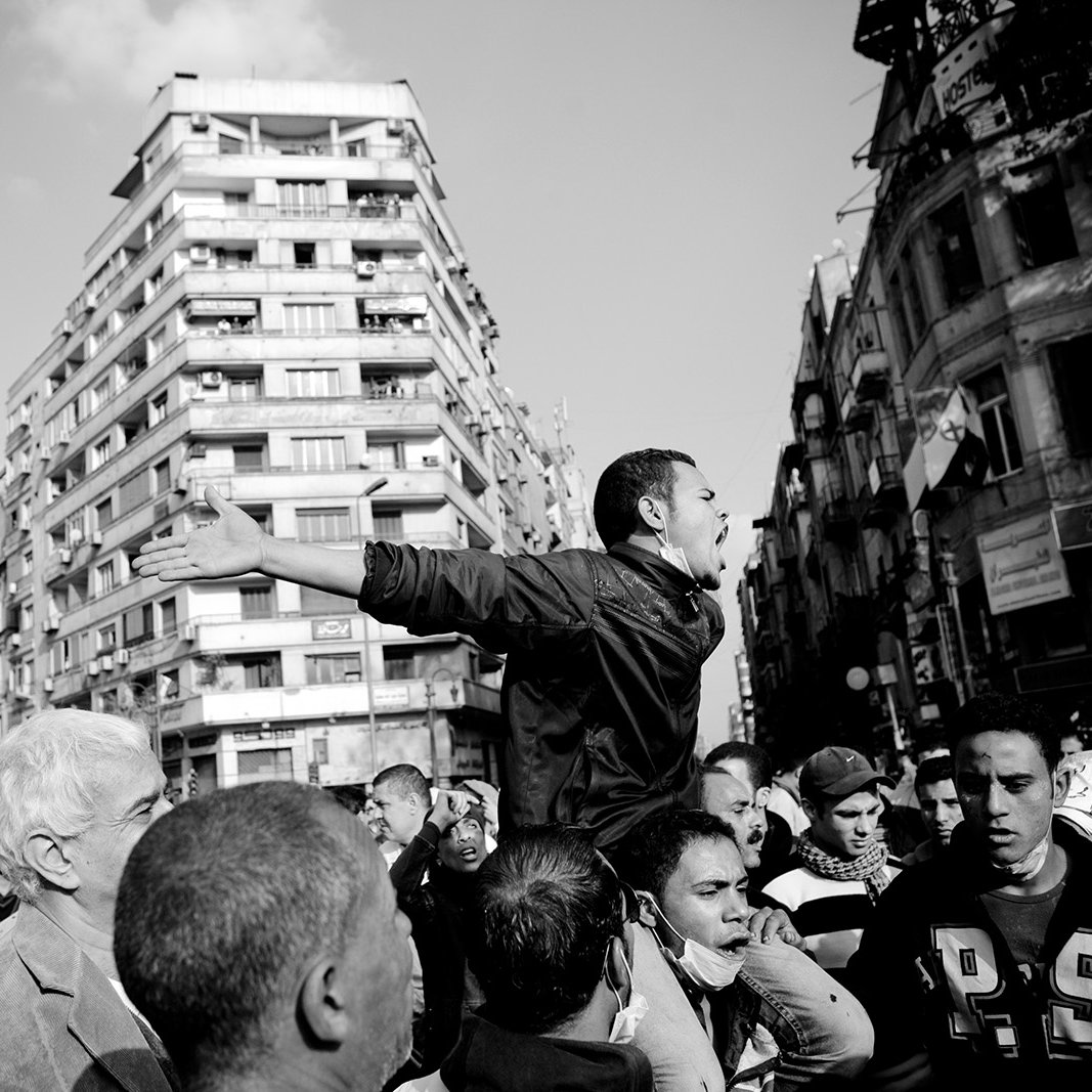 The image depicts a scene of a protest or demonstration. In the foreground, a young man is animatedly speaking or shouting, with his arms outstretched, indicating passion or urgency. Surrounding him are several people, some looking up at him with expressions of engagement or concern. The background features tall buildings, hinting at an urban environment. The photograph is in black and white, which emphasizes the intensity and emotions of the moment. The overall atmosphere conveys a sense of solidarity and activism.