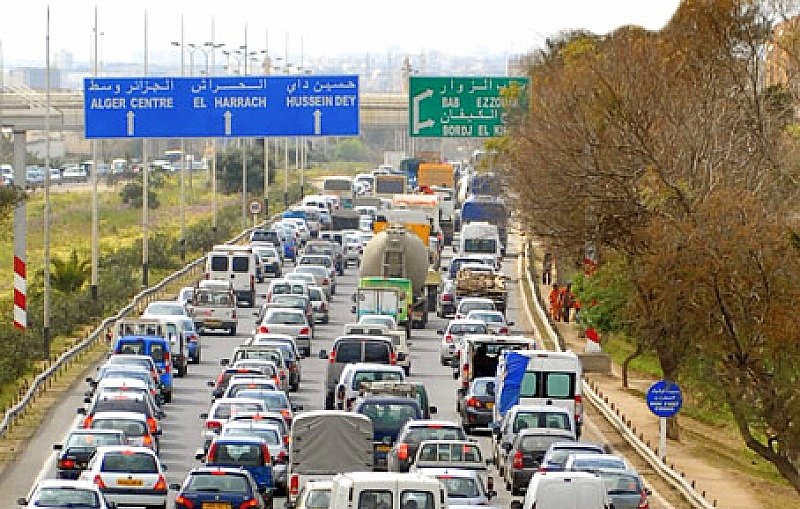 L'image montre une route très fréquentée avec une circulation dense. On peut voir de nombreux véhicules, y compris des voitures et des camionnettes, alignés les uns derrière les autres. Les panneaux de signalisation, écrits en arabe et en français, indiquent différentes destinations, comme "Alger Centre" et "Bab El Zawiya". Au loin, on aperçoit des arbres sur le bord de la route, ce qui suggère un environnement urbain. L'atmosphère semble être celle d'une journée ordinaire avec des bouchons routiers.