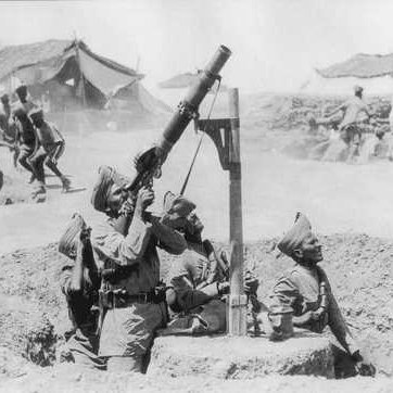 The image depicts a group of soldiers operating a mortar in a wartime setting. They are wearing military uniforms and are positioned in a trench-like area. One soldier is aiming the mortar, while other soldiers are present nearby, possibly providing support or preparing for an attack. In the background, there are tents suggesting a military camp, and additional soldiers can be seen, likely engaged in activity related to the ongoing conflict. The photo has a historical, black-and-white appearance, conveying a sense of the era in which it was taken.