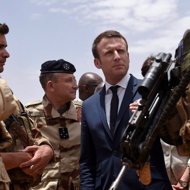 L'image montre un groupe de militaires en uniforme, dont certains portent des casques et des armes, rassemblés autour d'un homme en costume et cravate. Cet homme a une expression concentrée et semble en conversation avec les militaires, indiquant une interaction sérieuse. Le décor évoque un environnement militaire, probablement en extérieur, sous un ciel partiellement nuageux. Cette scène pourrait représenter une visite officielle ou une interaction avec des forces armées.