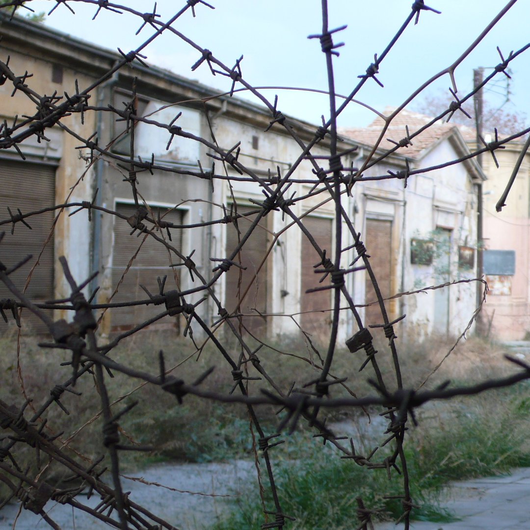 The image depicts an abandoned street scene, where several old buildings are visible with boarded-up windows and doors. The surroundings are overgrown with grass and weeds, suggesting neglect and disrepair. In the foreground, there are thorny branches or barbed wire, adding to the sense of desolation and possibly creating a feeling of confinement. The overall atmosphere feels eerie and forgotten, highlighting the passage of time in this once-active area.