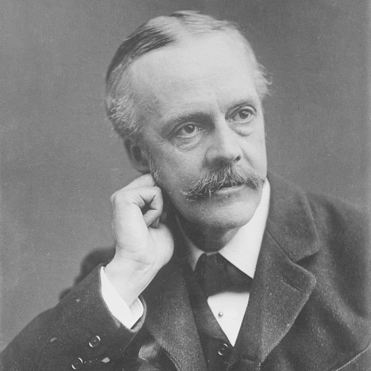 L'image montre un homme en costume vêtu d'une veste sombre et d'une cravate. Il a des moustaches bien entretenues et les cheveux légèrement grisonnants. Il pose en penchant légèrement la tête sur la main, affichant une expression pensive. Le fond est neutre, mettant en valeur son visage et son regard pensif.