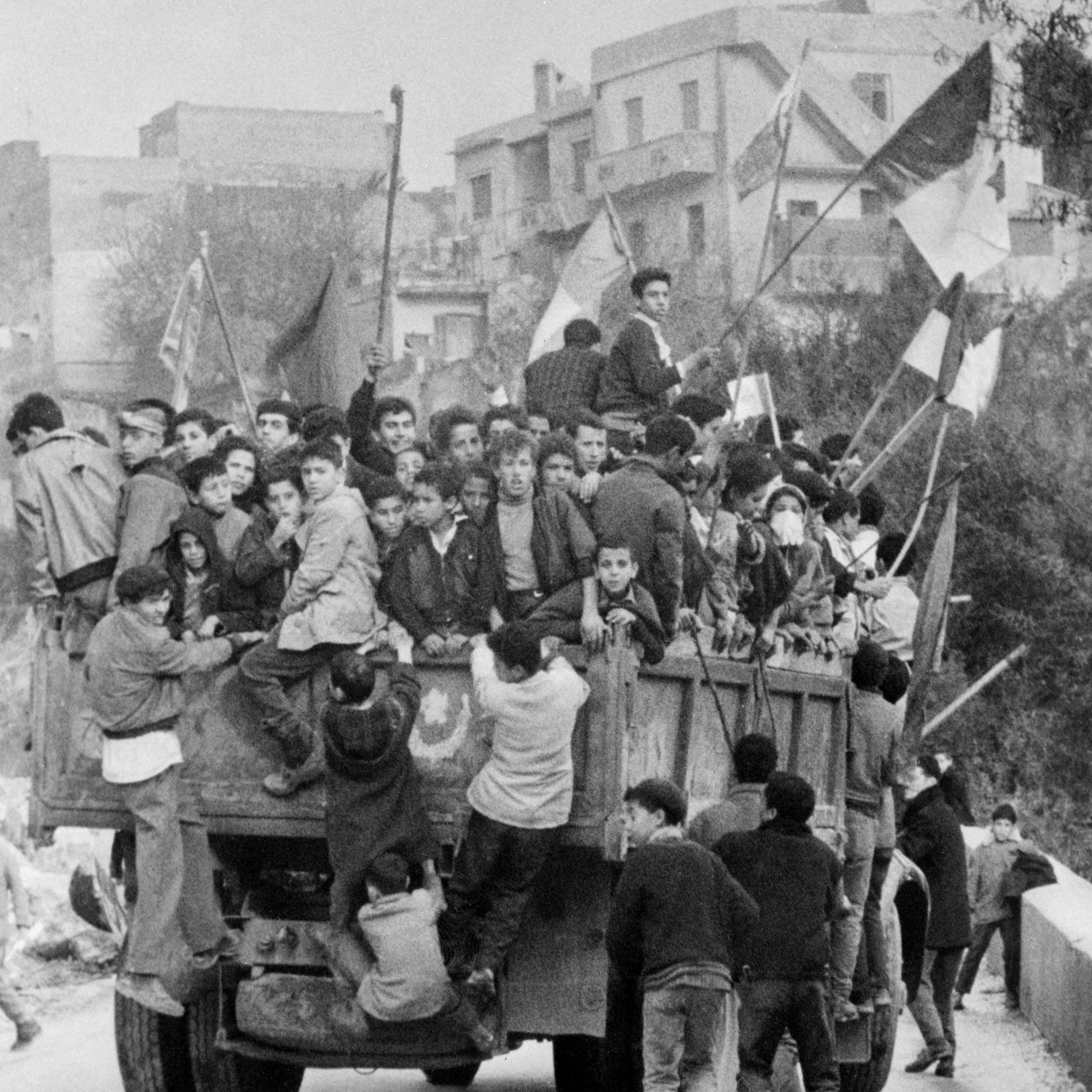 L'image montre un groupe de jeunes garçons et d'adolescents rassemblés sur un camion, entourés de drapeaux. Ils semblent célébrer un événement, peut-être une manifestation ou une fête. Les enfants sont penchés et accrochés au camion, reflétant un moment de camaraderie ou d'excitation collective. En arrière-plan, on aperçoit des bâtiments typiques d'une zone urbaine, contribuant à l'ambiance dynamique de la scène.