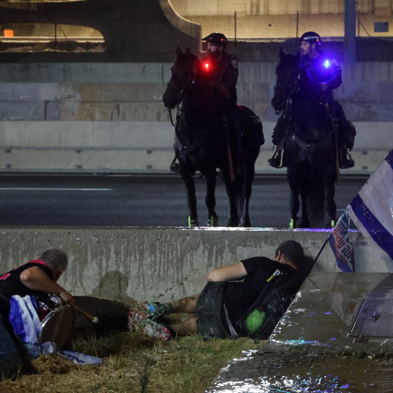 The image depicts a tense scene possibly related to a protest or civil unrest. In the foreground, two individuals appear to be on the ground, with one person lying down and another sitting nearby. They seem to be taking cover or seeking safety. In the background, two mounted police officers are visible, equipped with flashing lights on their helmets. There's also an Israeli flag positioned on the ground, suggesting that the situation might involve themes of national identity or political protest. The setting looks urban, with structures and concrete present, contributing to a sense of urgency in the atmosphere.