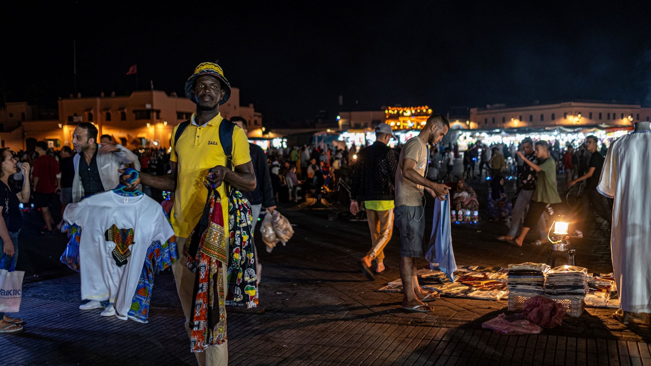 L'image montre un marché nocturne animé, probablement dans un endroit comme Marrakech. On peut voir des gens se déplacer dans une ambiance vibrante. Au premier plan, un homme porte un t-shirt jaune et tient plusieurs vêtements traditionnels colorés. En arrière-plan, d'autres personnes flânent et explorent les étals, tandis que des lumières scintillantes créent une atmosphère festive. Des tapis et d'autres articles sont visibles au sol, ajoutant à l'attrait du marché.